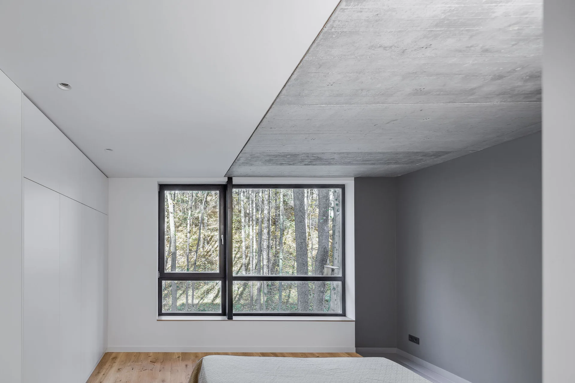 A minimalist bedroom features wood flooring, flat-front white wardrobes, and a split concrete ceiling next to a large window overlooking a forest.