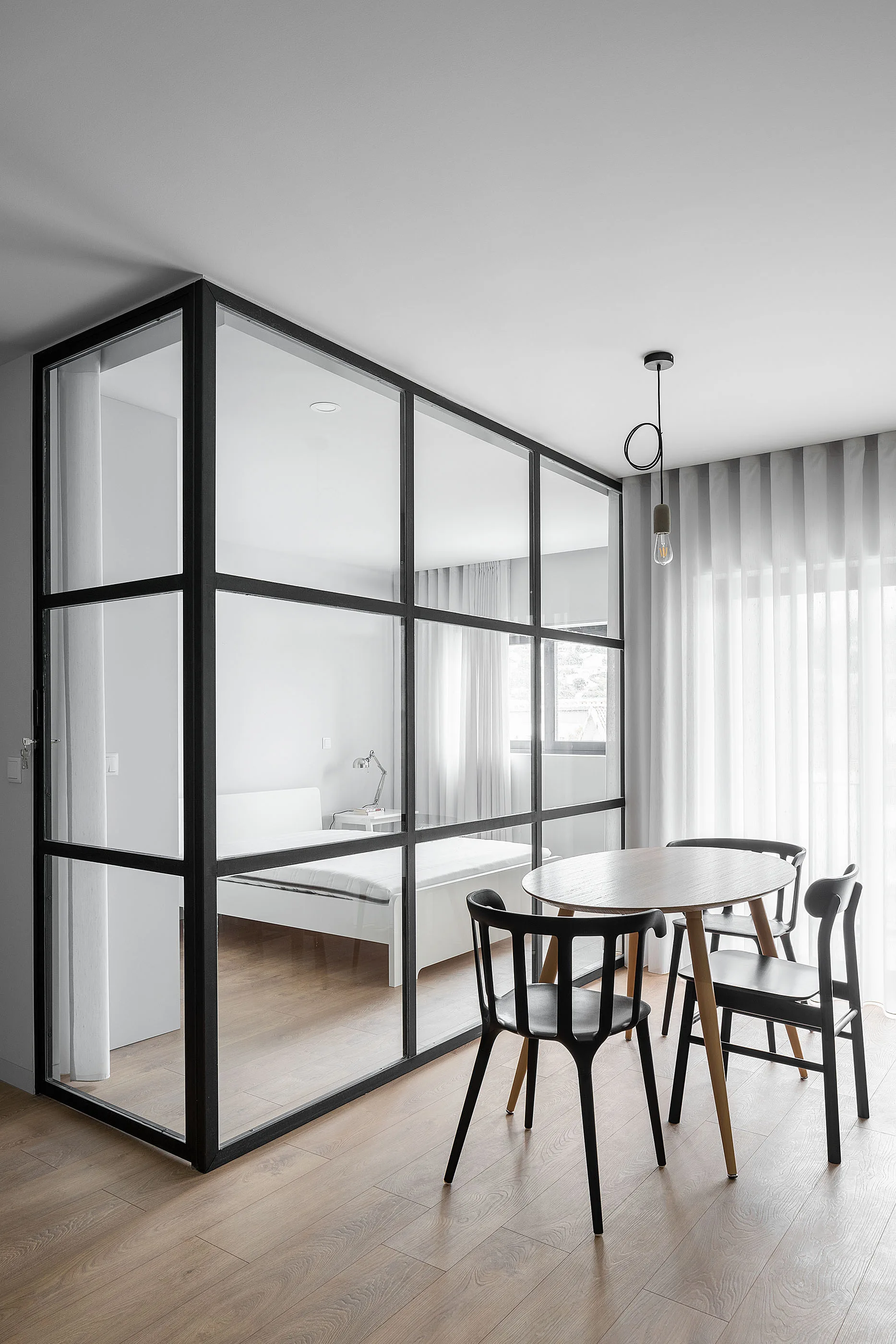 A round wood table and black chairs sit next to a black-framed glass wall partition separating a dining area from a bedroom.