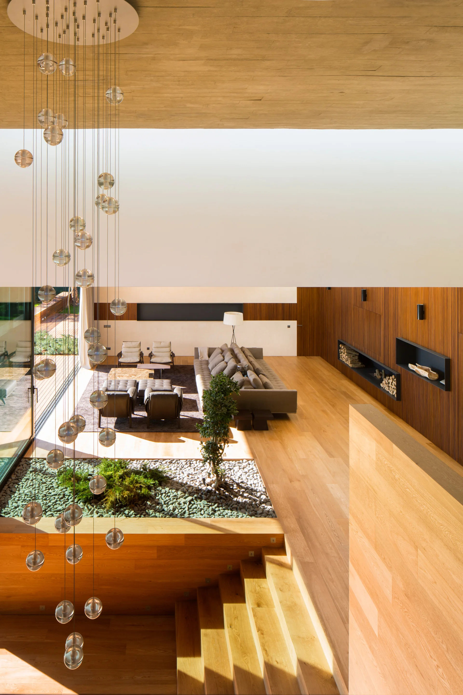 An overhead view of a modern living room featuring a grey sectional sofa, an indoor sunken rock garden, a staircase, and a tall glass chandelier.