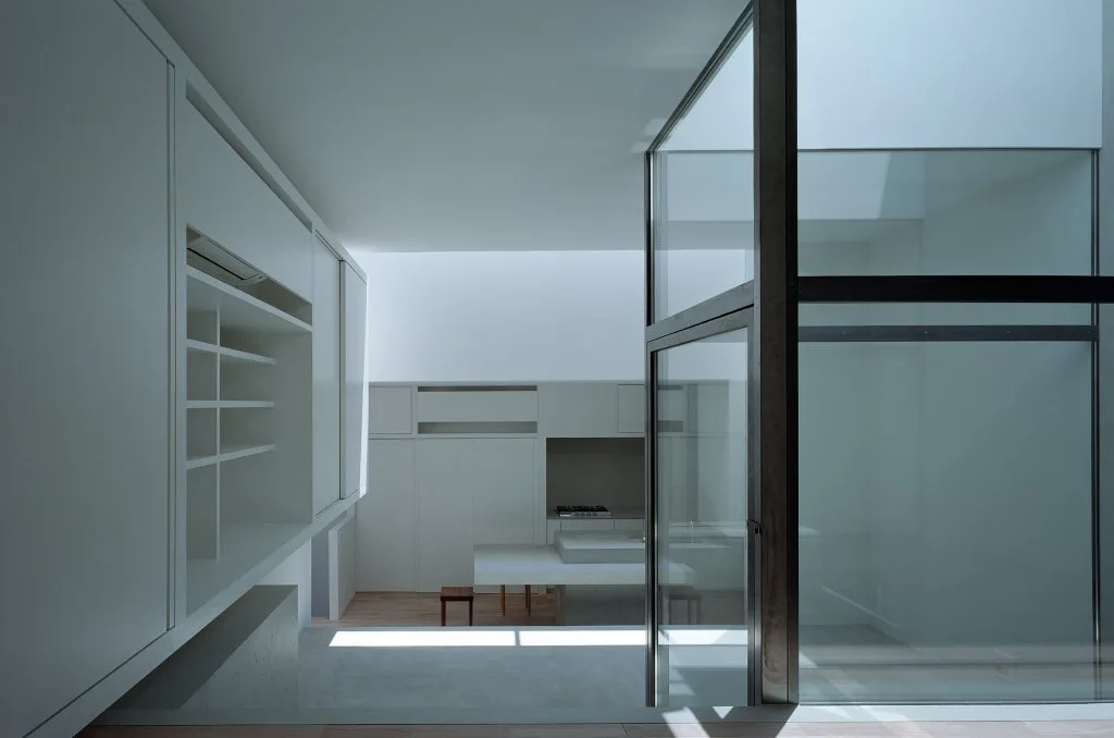 A minimalist white kitchen features handle-less cabinetry, a stone island, and a large steel-framed glass partition that draws in natural light.