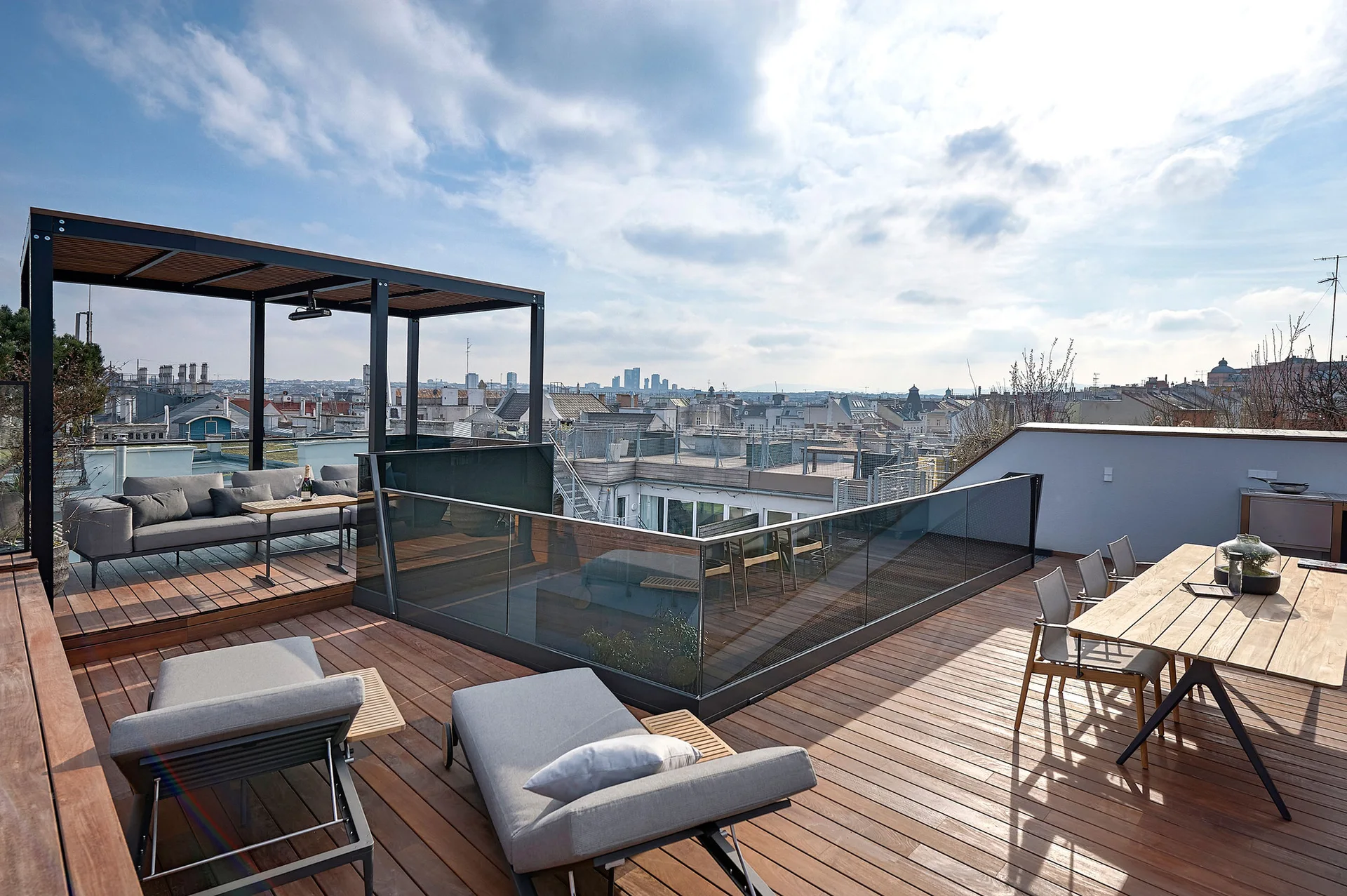 A spacious rooftop terrace features a wooden plank deck, grey lounge chairs, a dining table, and a black metal pergola under a cloudy sky.