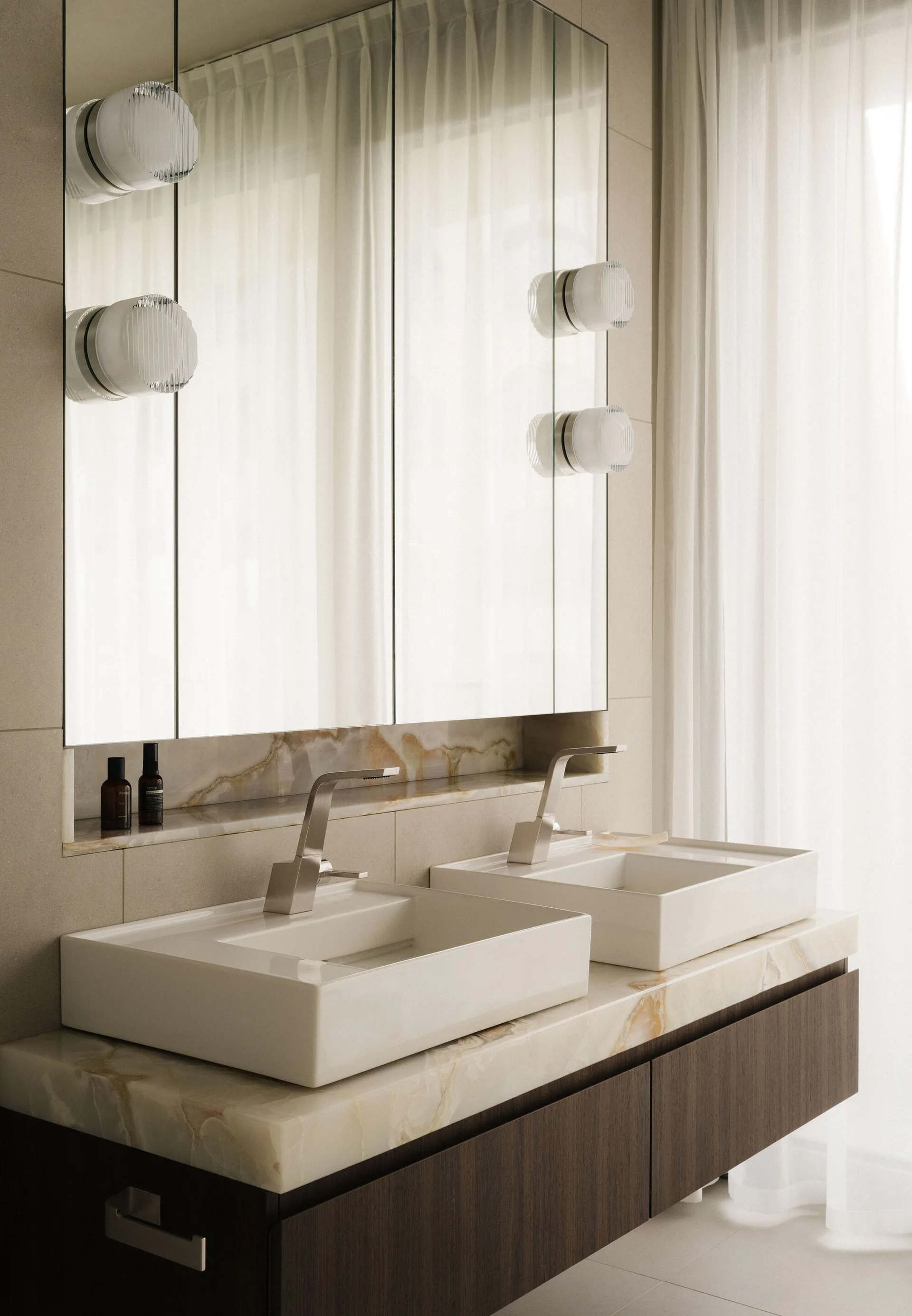 Two white vessel sinks rest on a veined stone vanity countertop beneath mirrored medicine cabinets with integrated glass sconces.