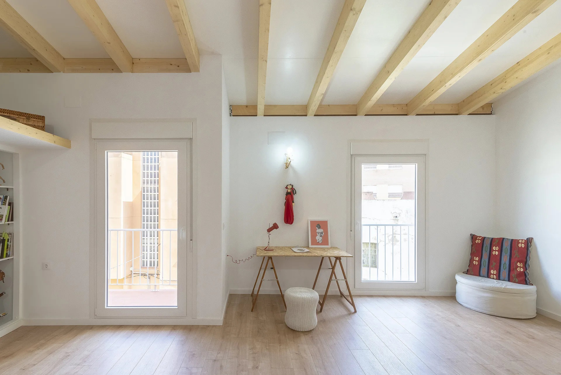 A bright room with exposed light wood ceiling beams, white walls, a trestle desk with a red lamp, and two glass patio doors.