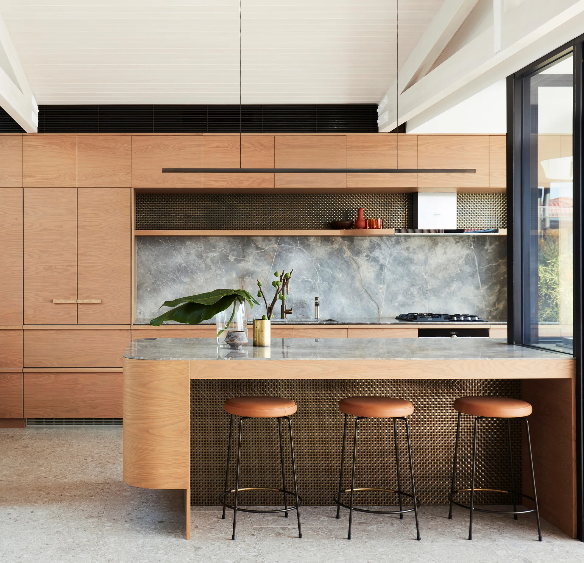 A modern kitchen with wood cabinetry, a stone-topped island featuring a curved wood end, bronze mesh paneling, and three leather bar stools.