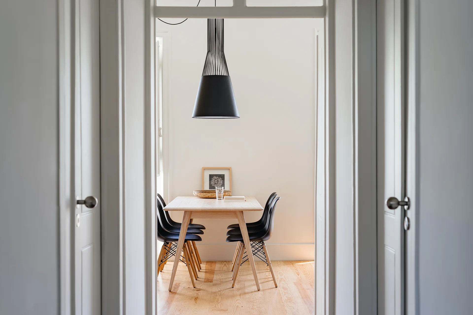 A view through a white doorway reveals a minimalist dining room featuring a light wood table, four black chairs, and a black pendant light.