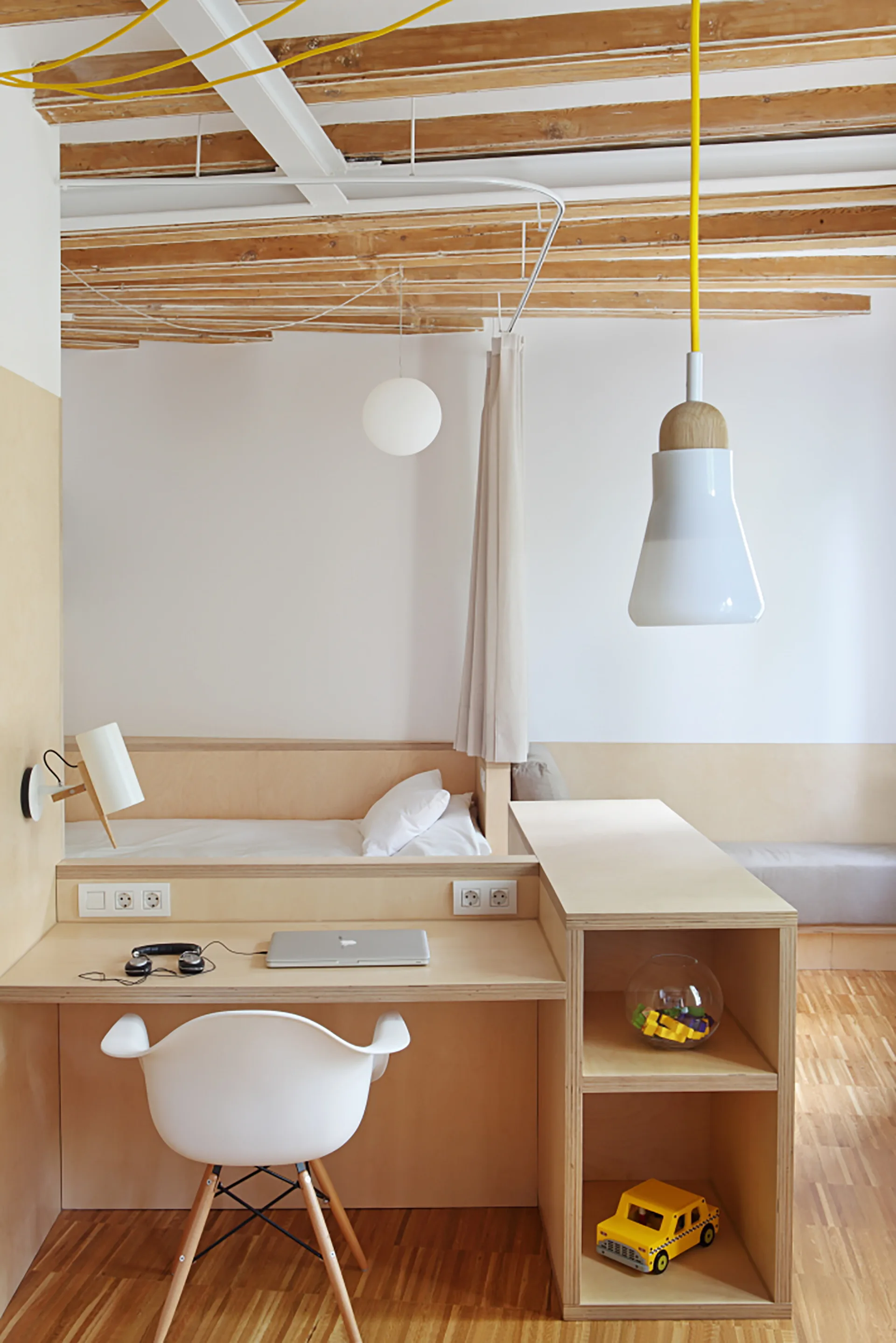 A compact minimalist bedroom features a built-in plywood desk with a white chair, integrated shelving, a bed, and exposed wooden ceiling beams.