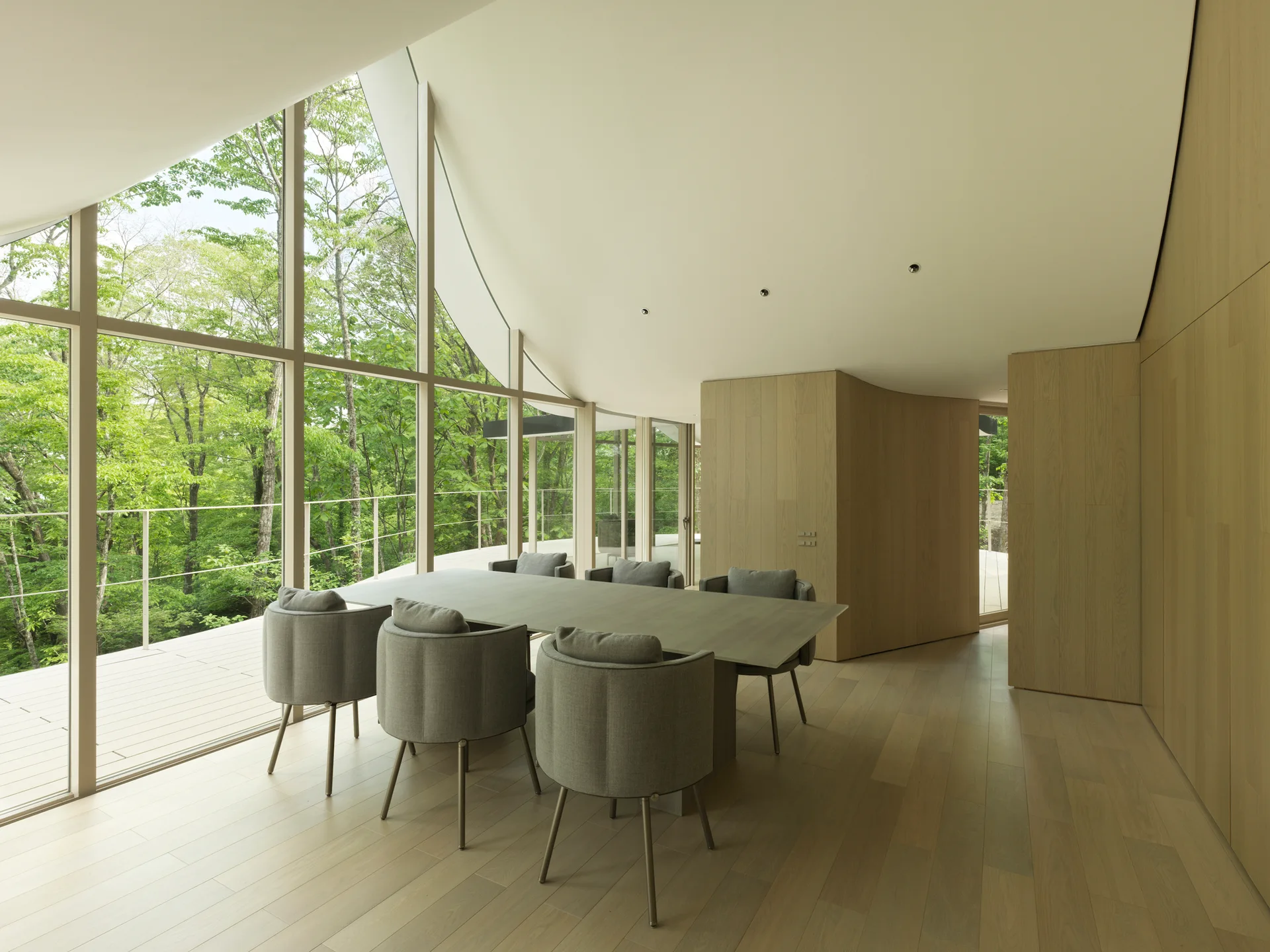 A minimalist dining table with grey chairs sits next to a large glass window wall showing a green forest under a curved ceiling.