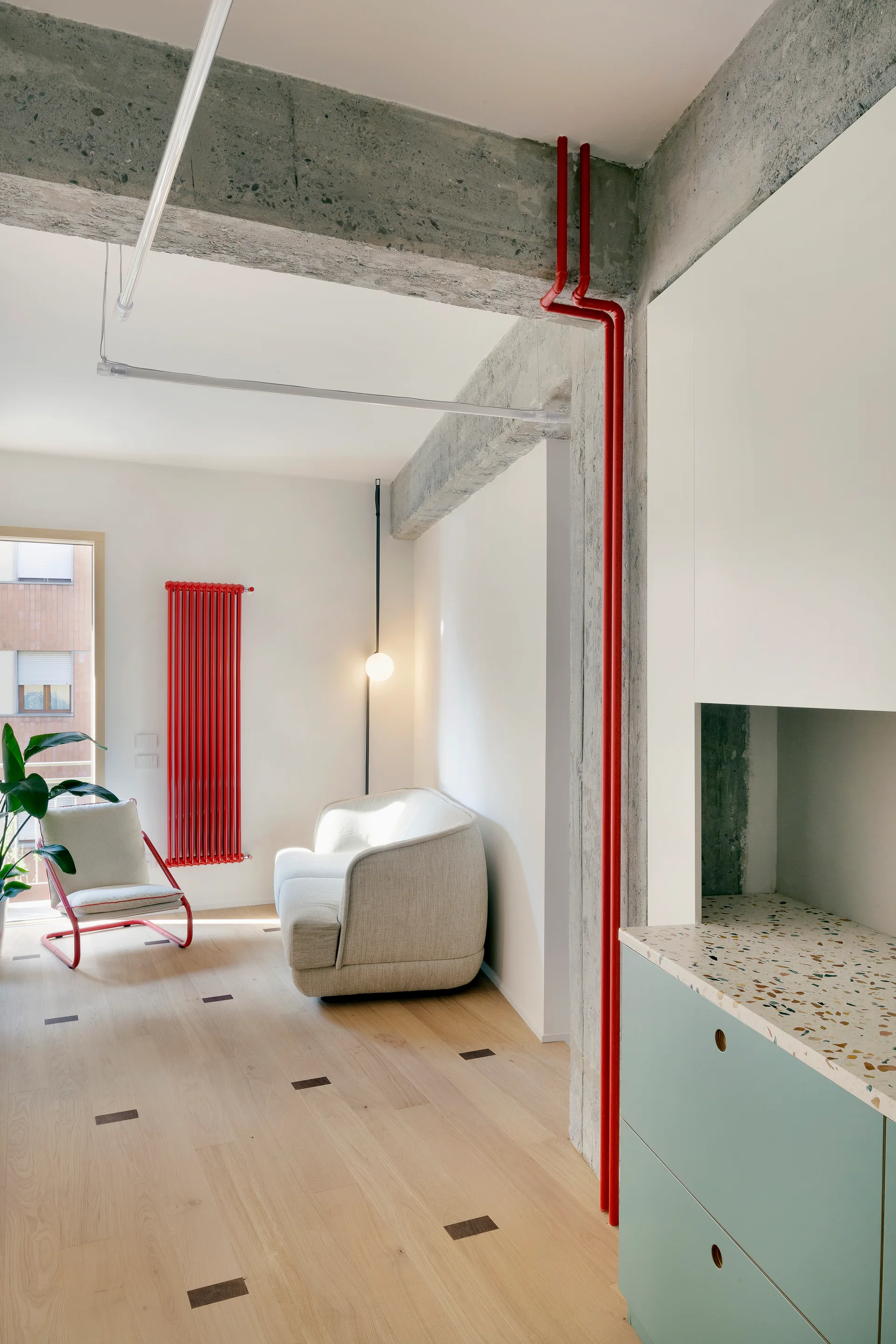 A modern minimalist living room with a beige sofa, exposed concrete beams, and red plumbing pipes along the wall.