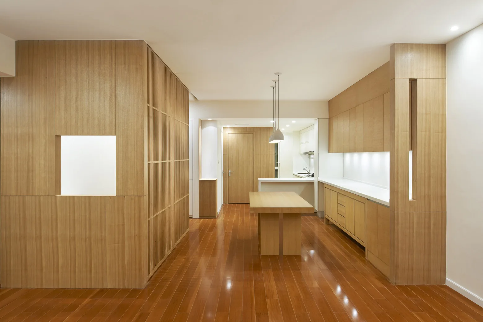 A minimalist kitchen featuring light wood cabinetry, a central dining table, and a vertical wood slat partition wall on polished hardwood floors.