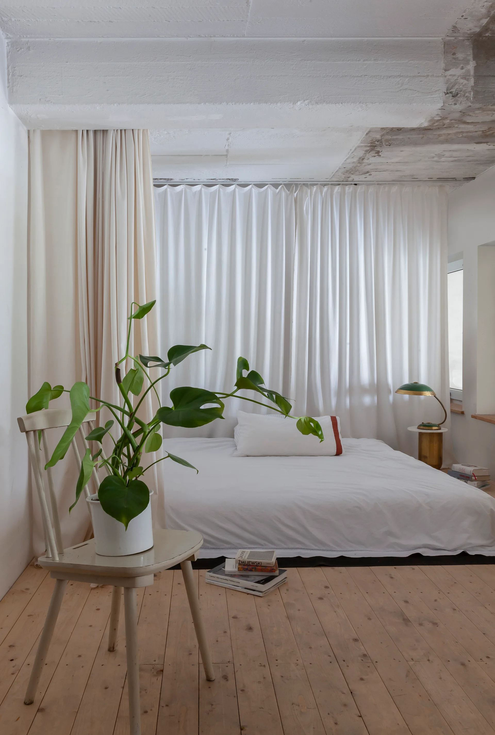 A minimalist bedroom features a low bed against white floor-to-ceiling curtains, light wood floors, and a green plant on a chair.