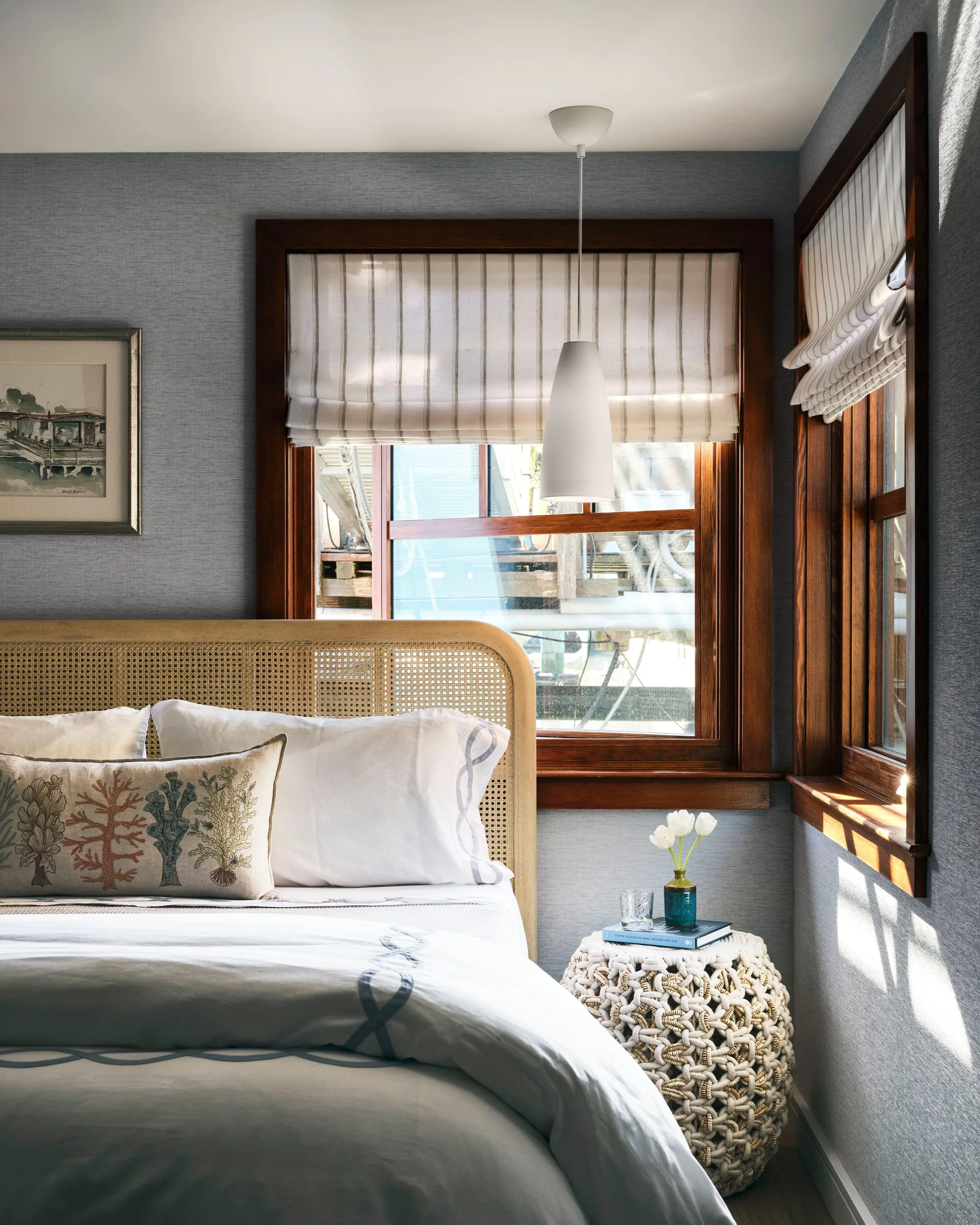 A cozy bedroom corner features a cane headboard, striped Roman shades inside dark wood window frames, and a woven rope nightstand.