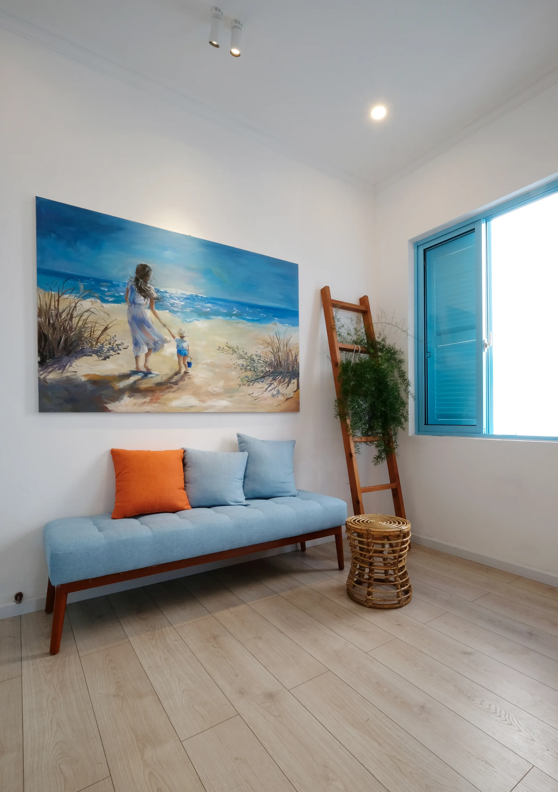 A light blue upholstered bench with colorful pillows sits beneath a beach painting next to a decorative wooden ladder and blue window.
