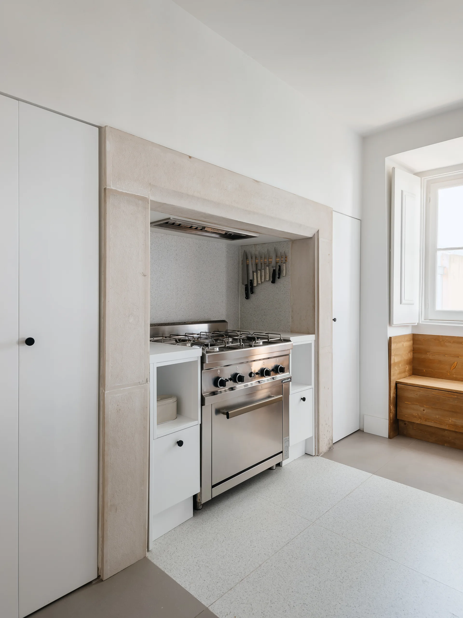 A modern kitchen features a stainless steel range stove recessed in a limestone alcove next to white minimalist cabinets and a wooden window bench.