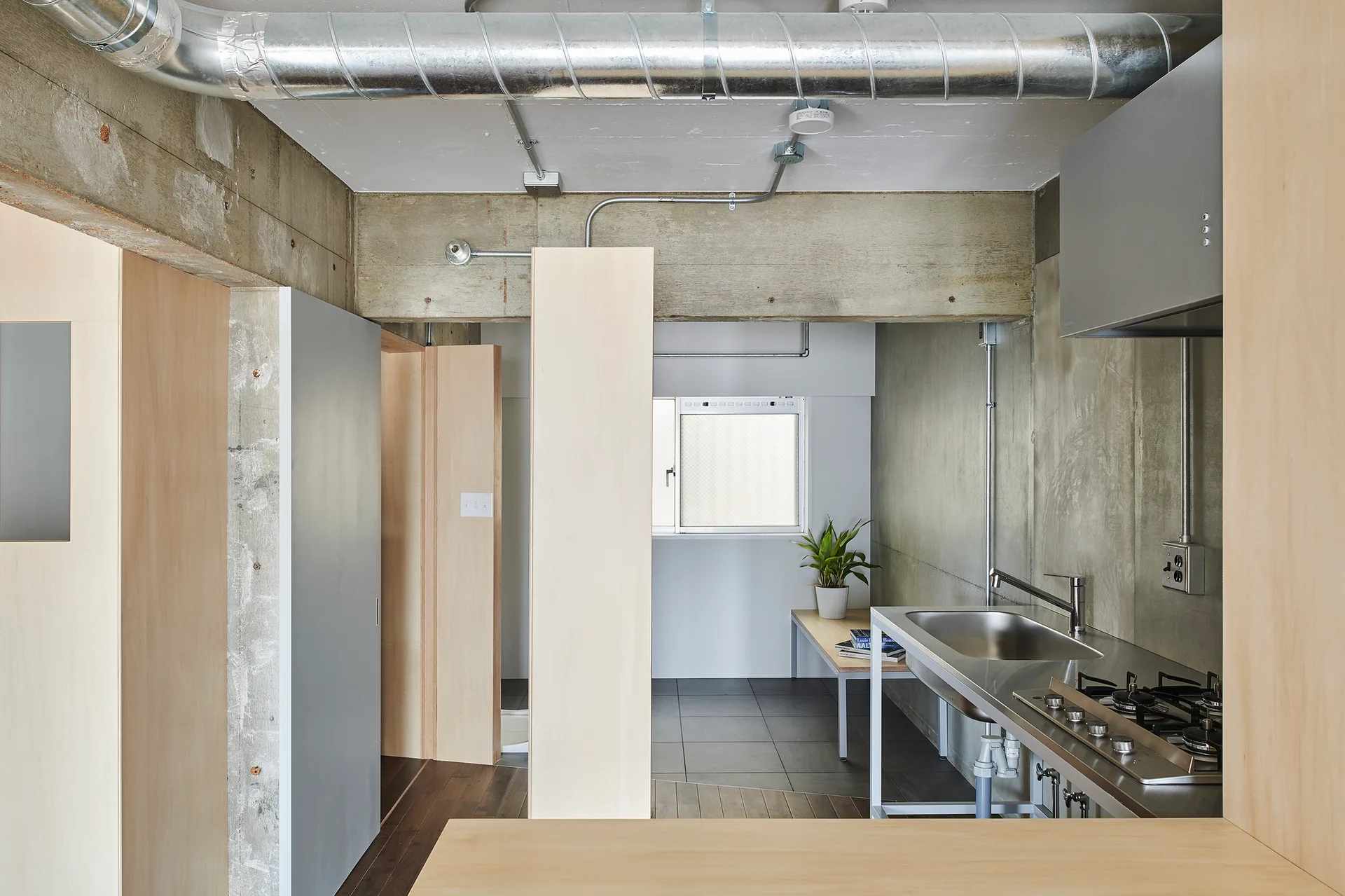 An industrial kitchen featuring raw concrete walls, warm plywood partitions, exposed silver ceiling ductwork, and a stainless steel sink unit.
