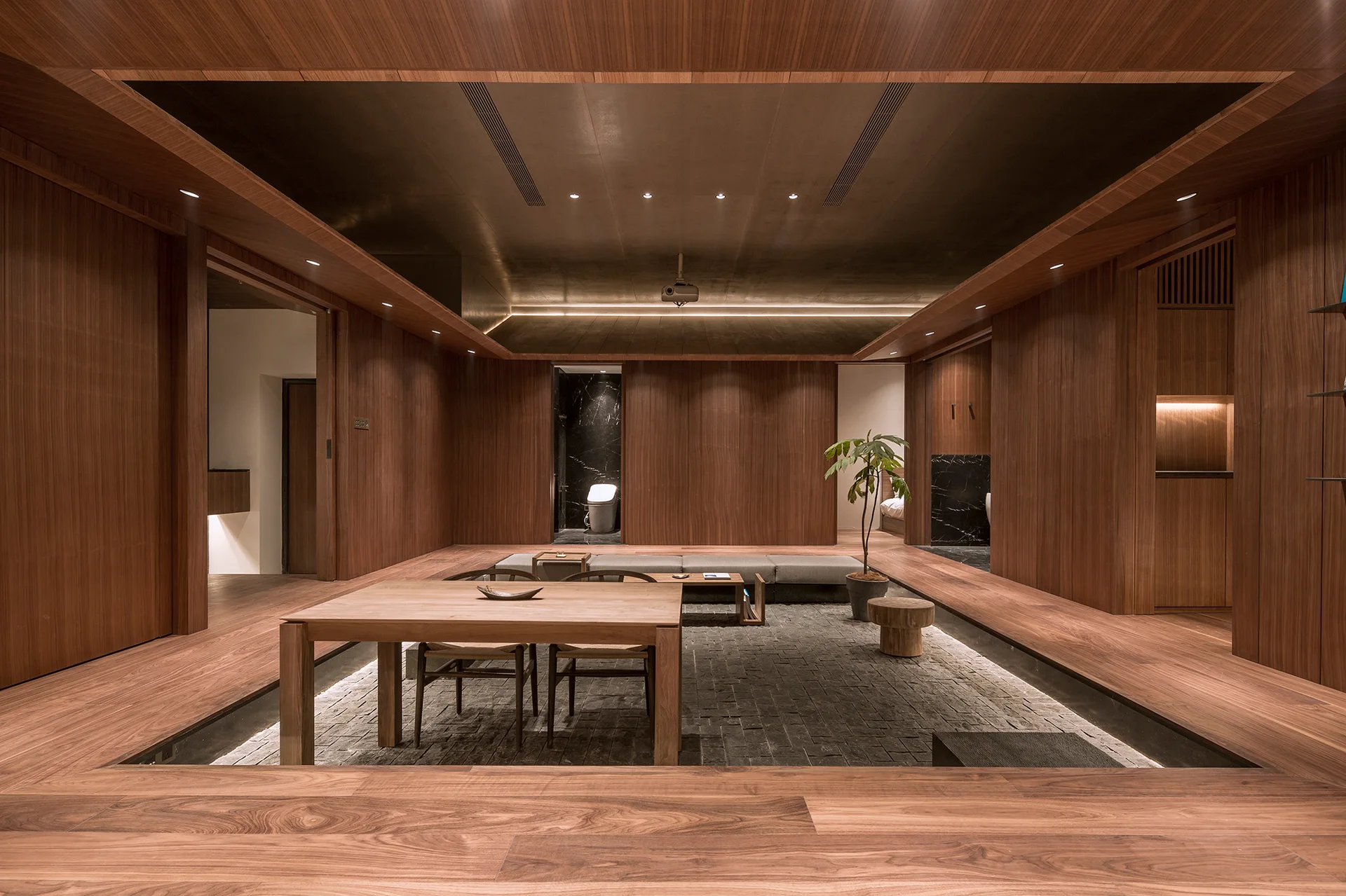 A minimalist room with wood-paneled walls and ceiling features a sunken living area with grey stone flooring and low-profile furniture.