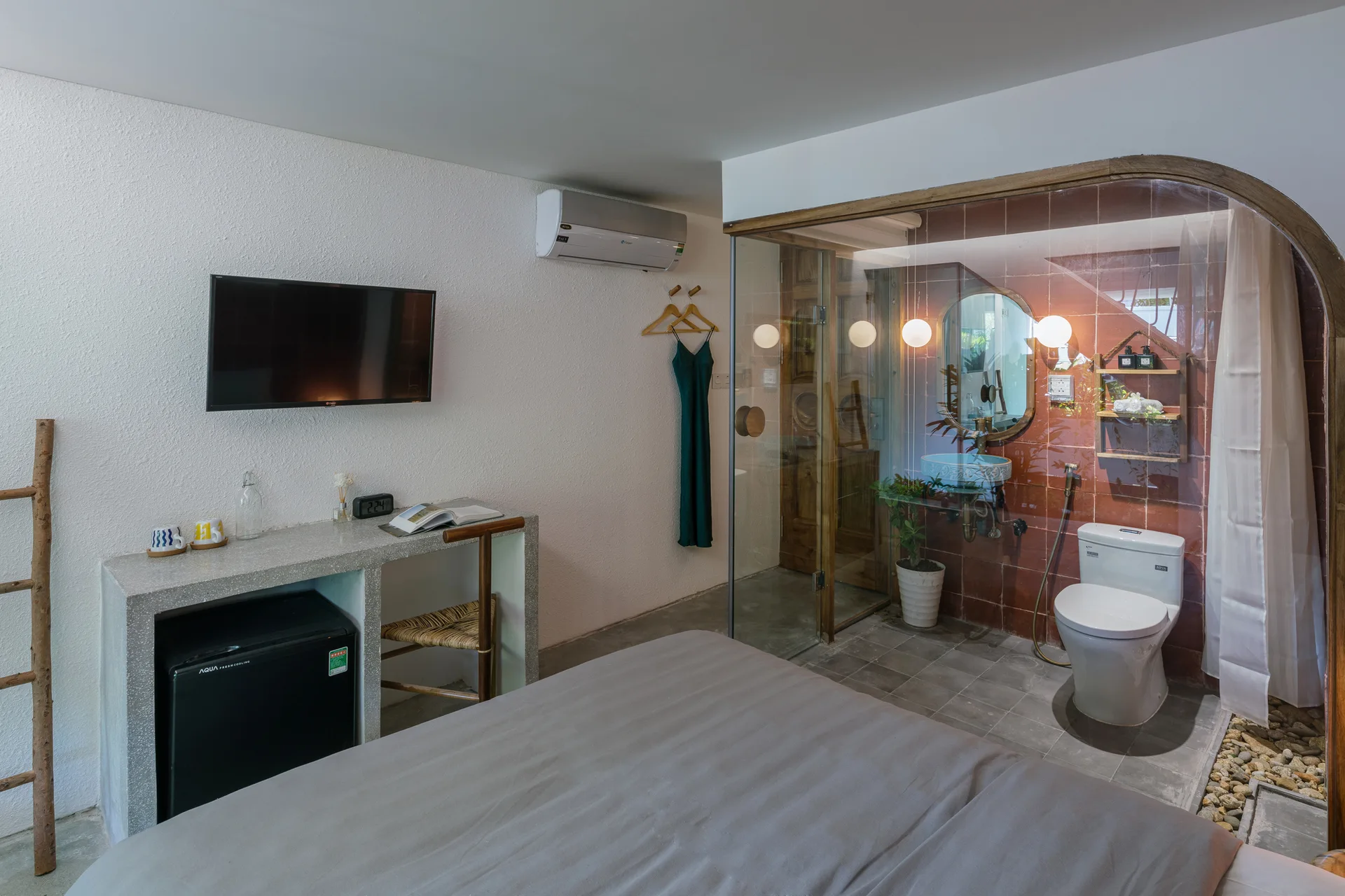 A minimalist bedroom featuring a concrete desk with a mini fridge and a glass-enclosed ensuite bathroom tiled in terracotta.