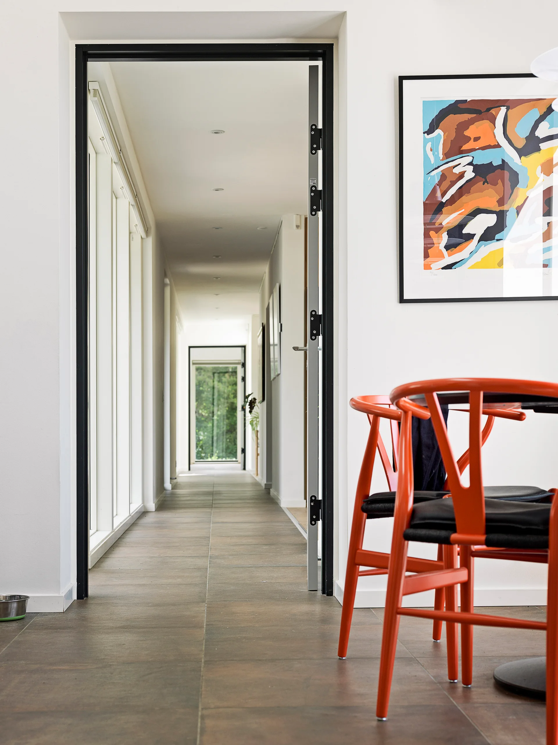 A view looking down a long white hallway through a black-framed doorway, with bright orange wishbone-style chairs and framed art in the foreground.