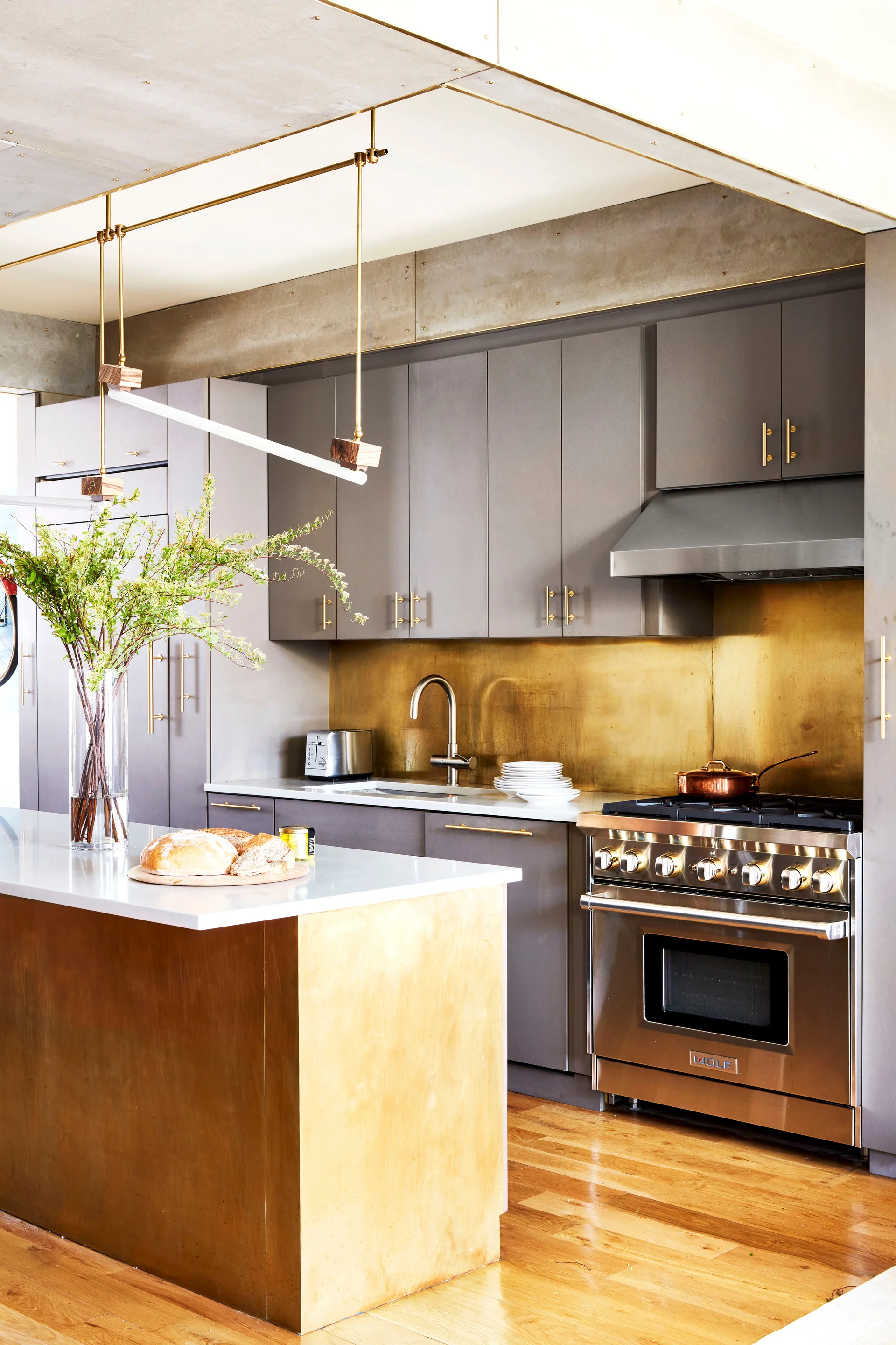 A modern kitchen featuring matte gray cabinets, a solid brass backsplash, a brass-clad island, and an exposed concrete ceiling.