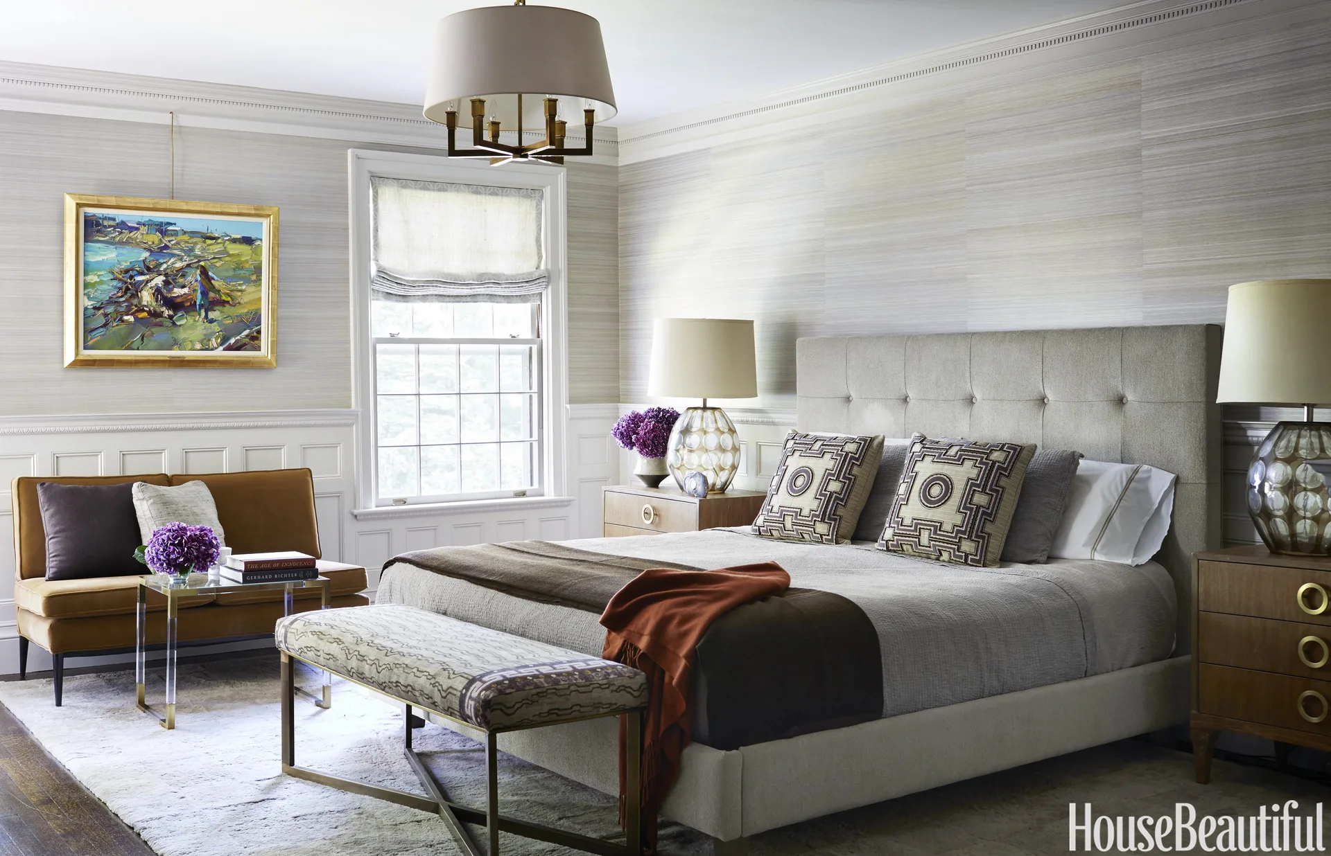 A transitional bedroom featuring an upholstered bed, wooden nightstands with large glass lamps, a window, and a small gold-toned sofa.