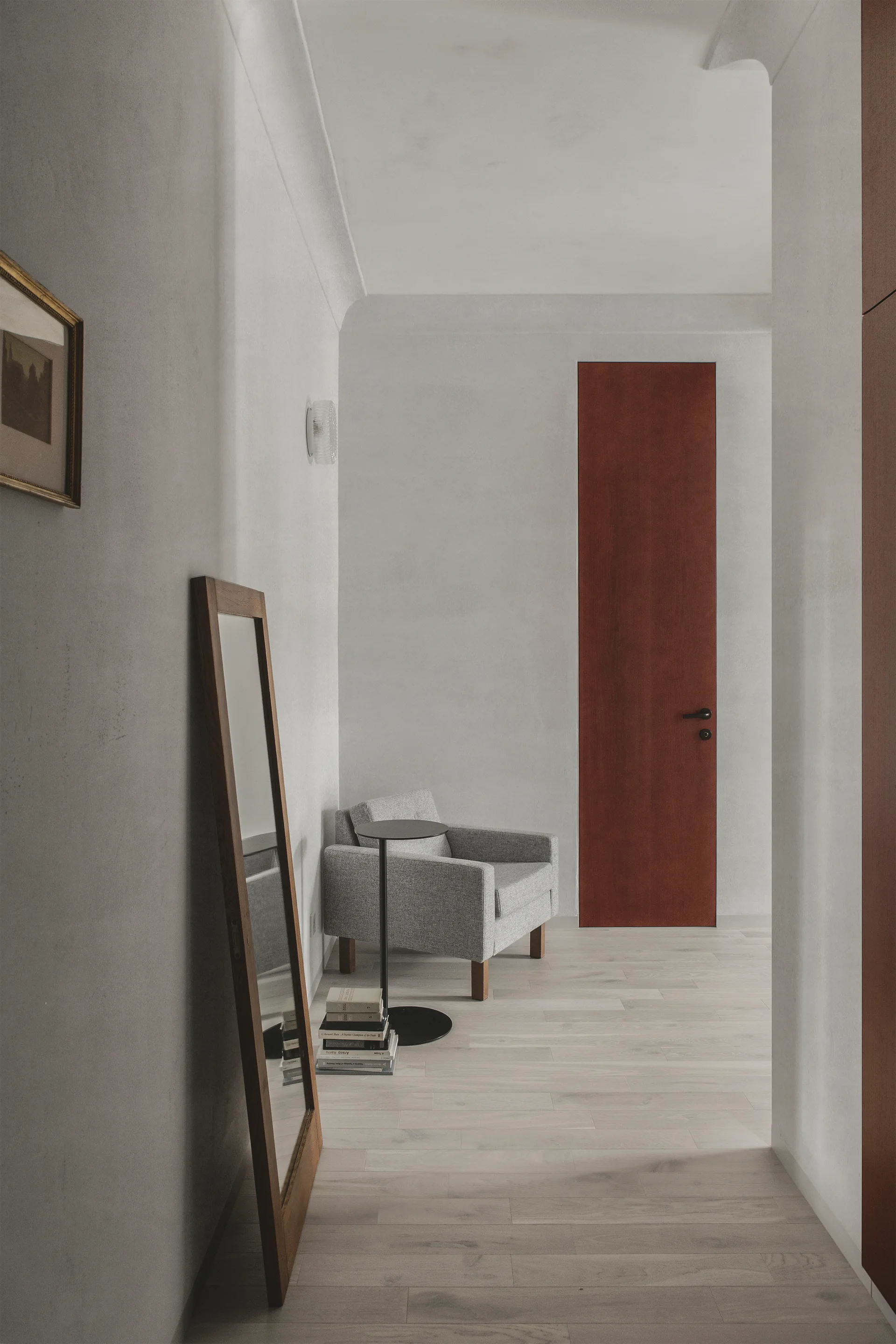 A minimalist hallway featuring light wood floors, a grey armchair, a leaning wooden floor mirror, and a tall reddish-brown wood door.