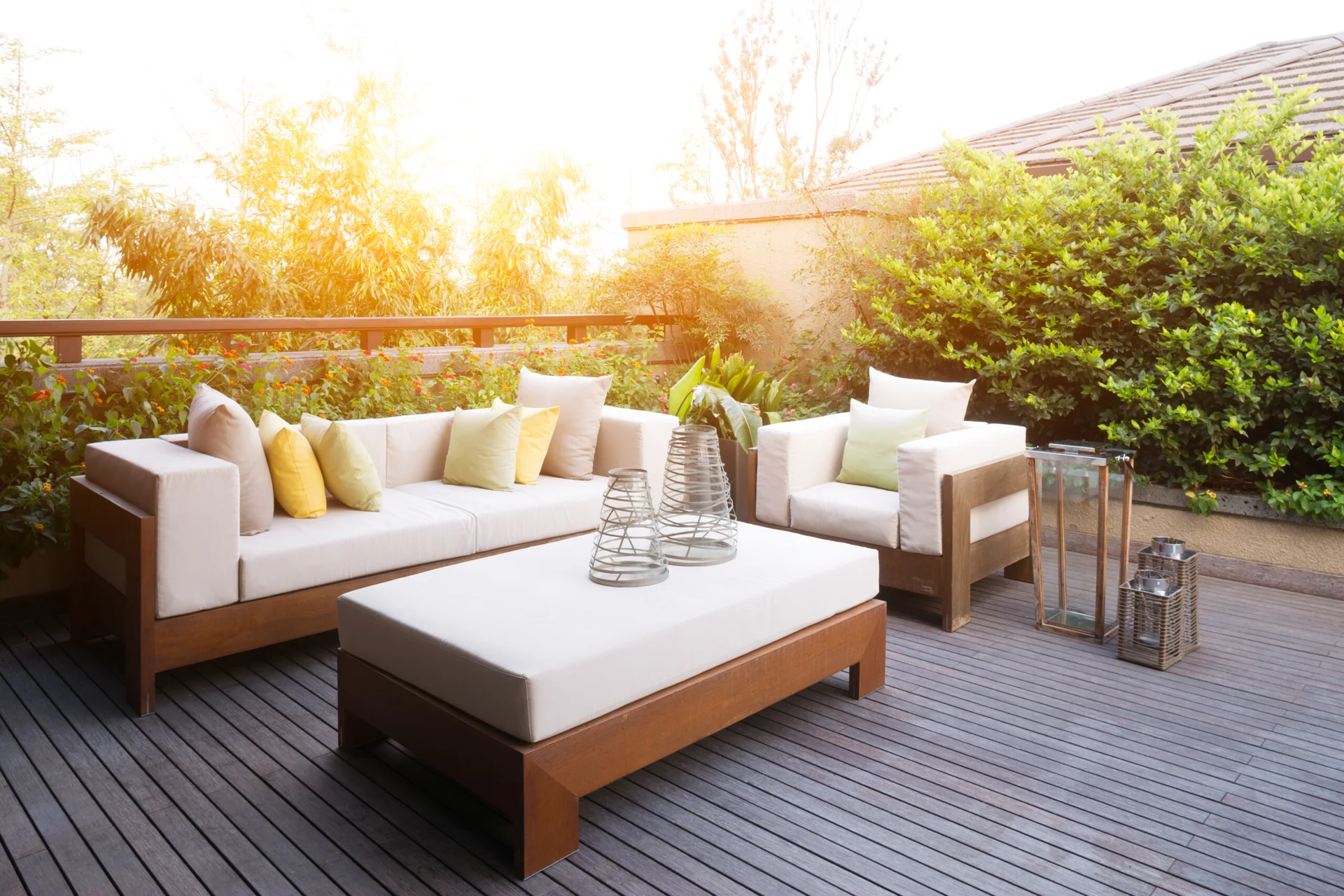 A wooden deck terrace features a beige sofa and matching armchair, styled with yellow throw pillows, wire decorative vessels, and potted greenery.