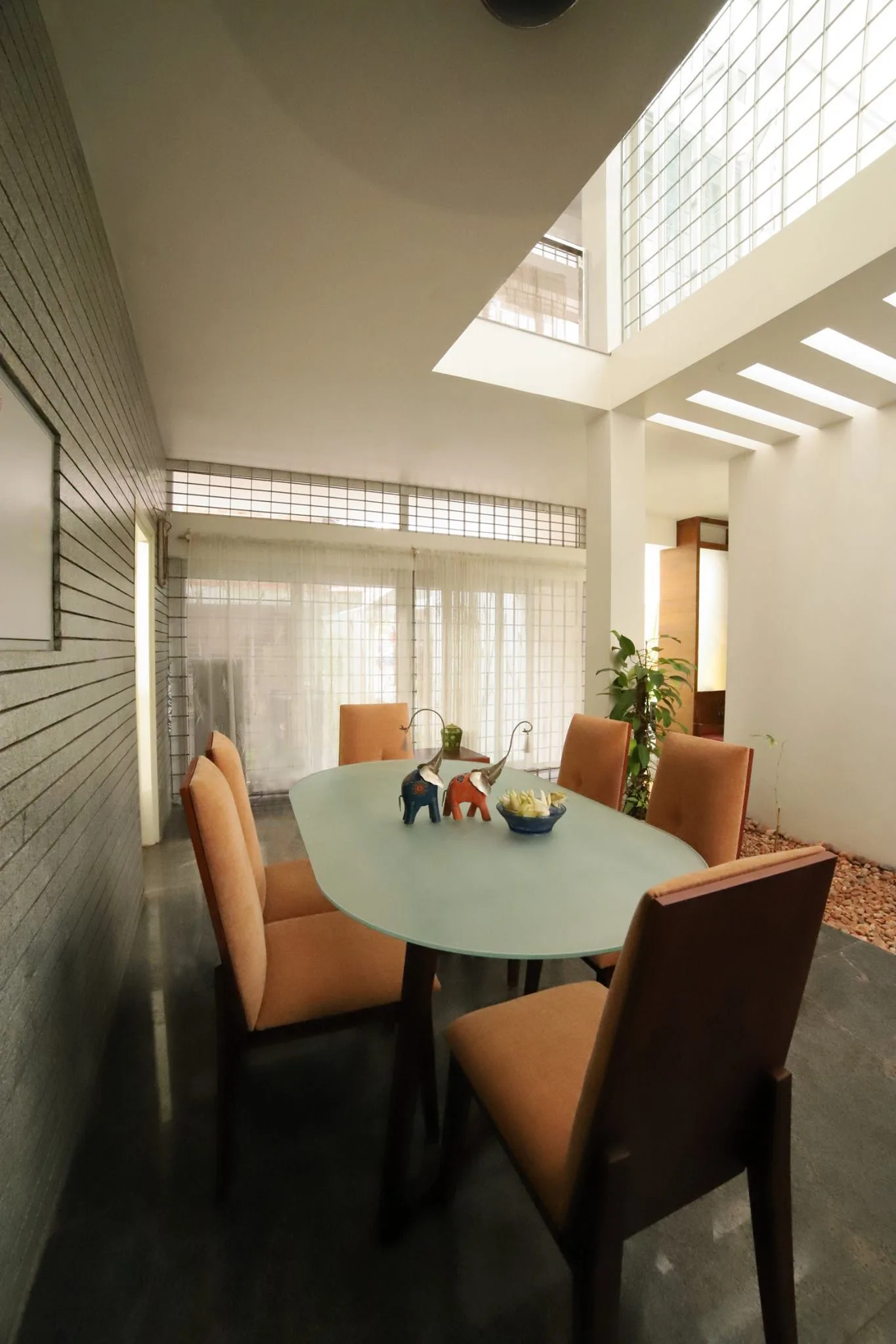 An oval glass dining table and orange upholstered chairs sit beneath a high skylight in a room with a slatted gray accent wall.