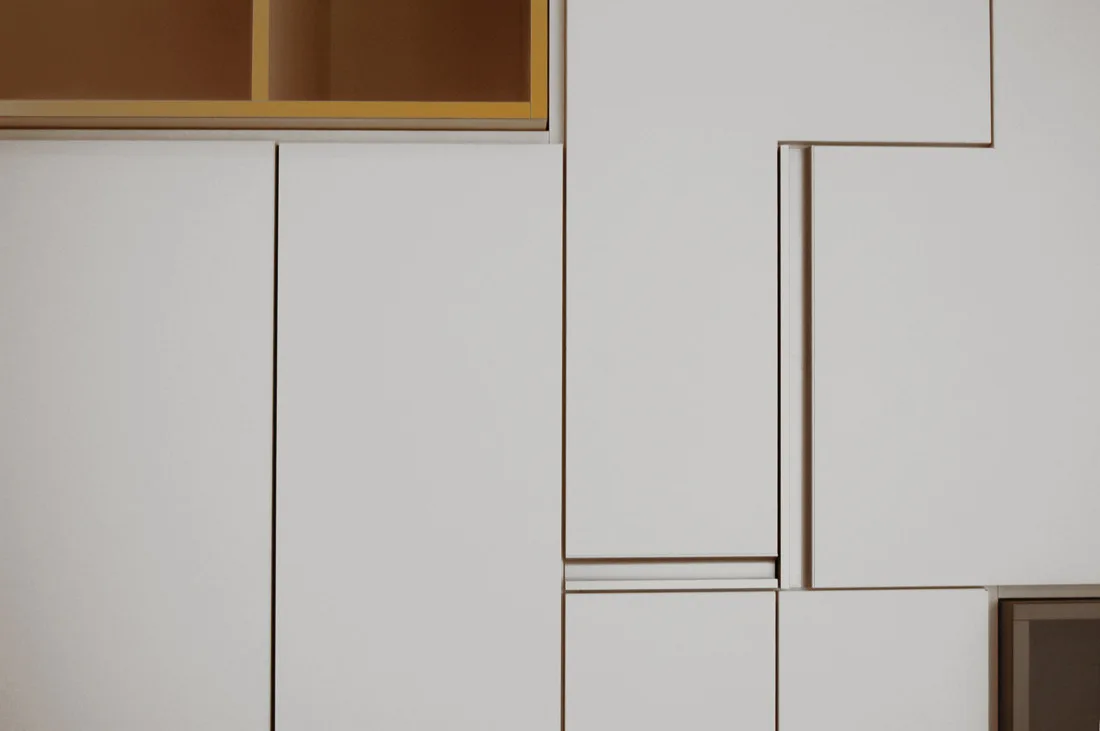 Matte white cabinet doors feature an asymmetrical geometric layout with integrated recessed handles and a gold open-shelf accent.