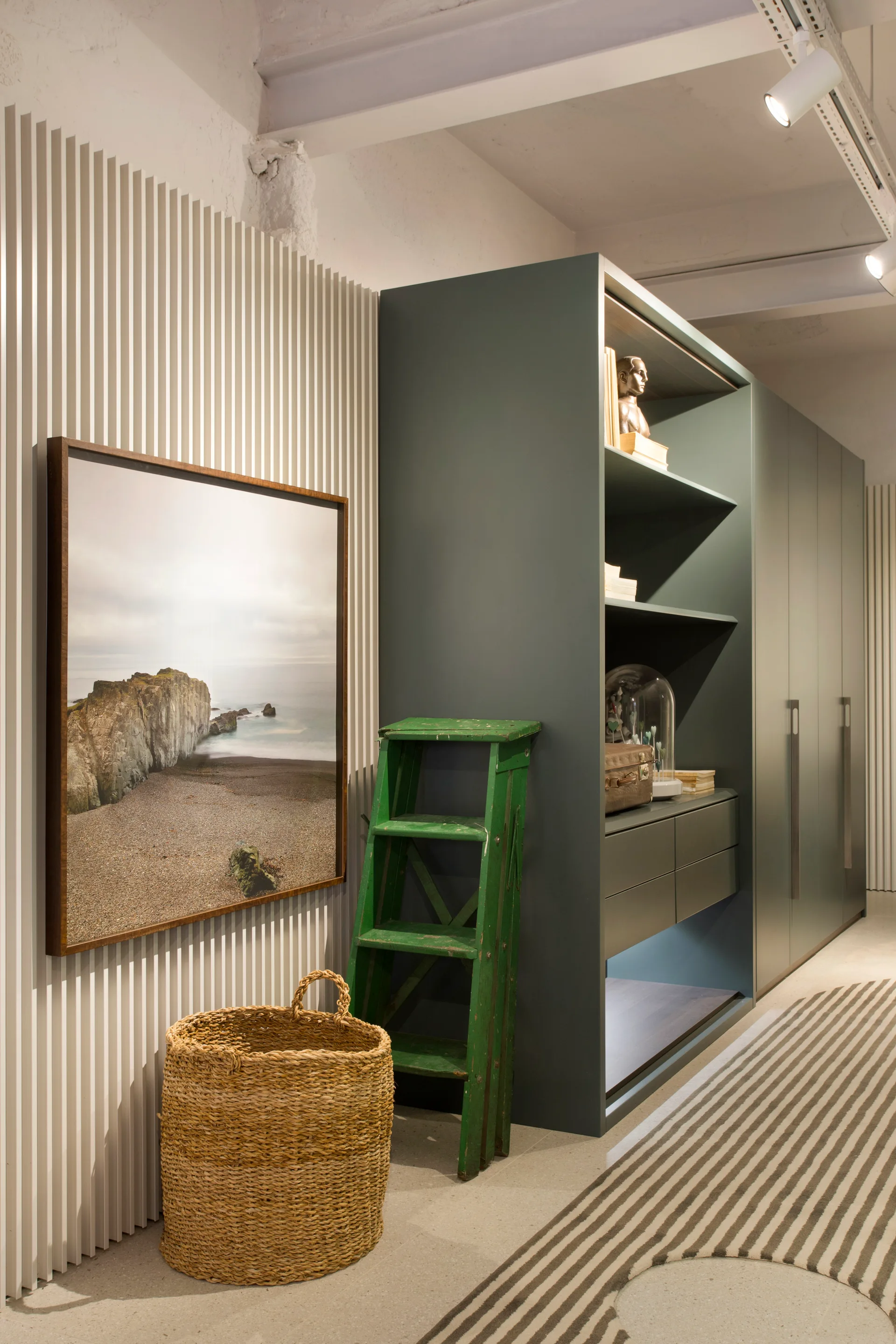 A dark green custom cabinet stands next to an off-white vertical slatted wall featuring framed artwork, a green wooden step ladder, and a woven basket.