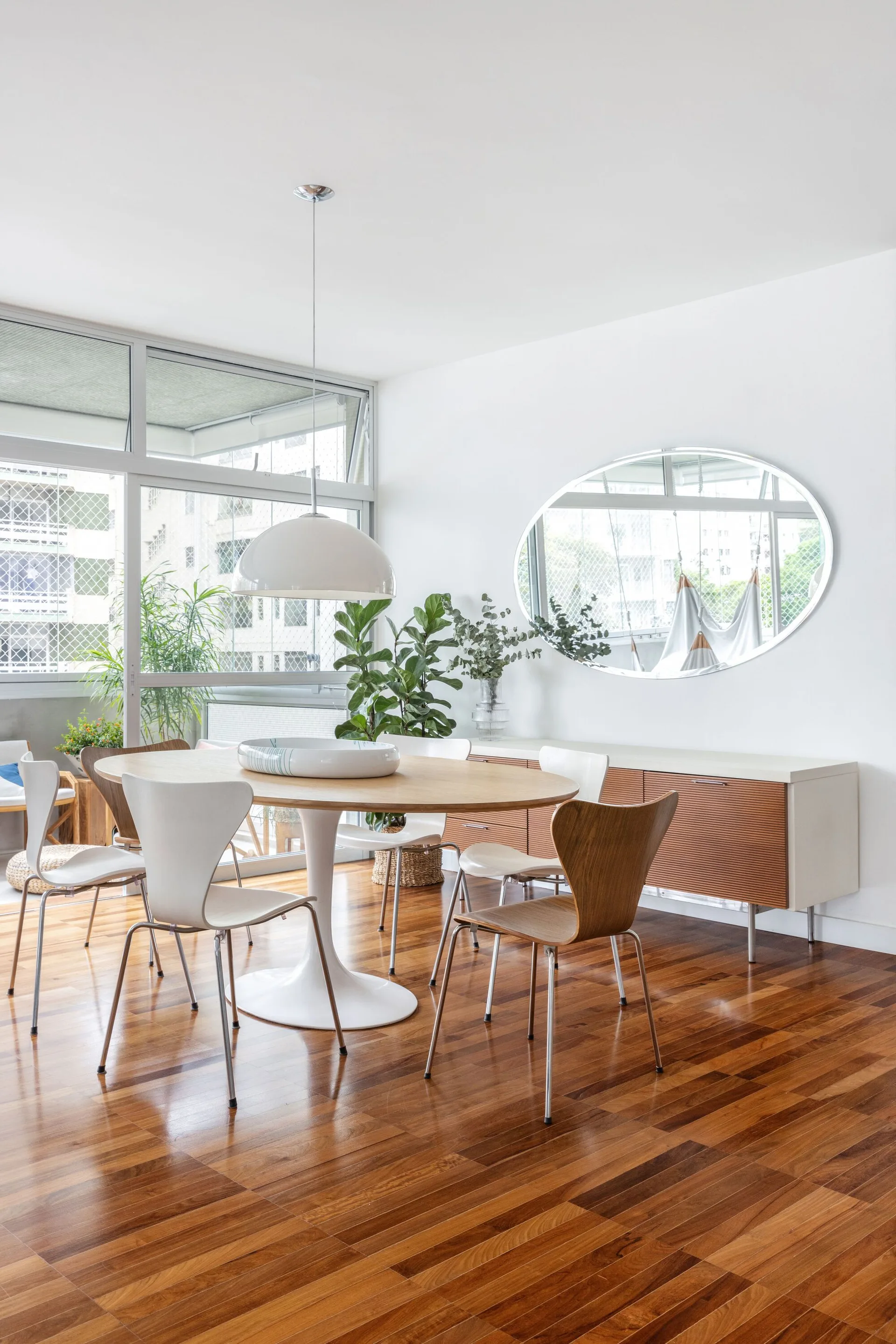 A bright dining room featuring an oval wood table, mixed chairs, a white dome pendant light, and a slatted sideboard beneath a large oval mirror.