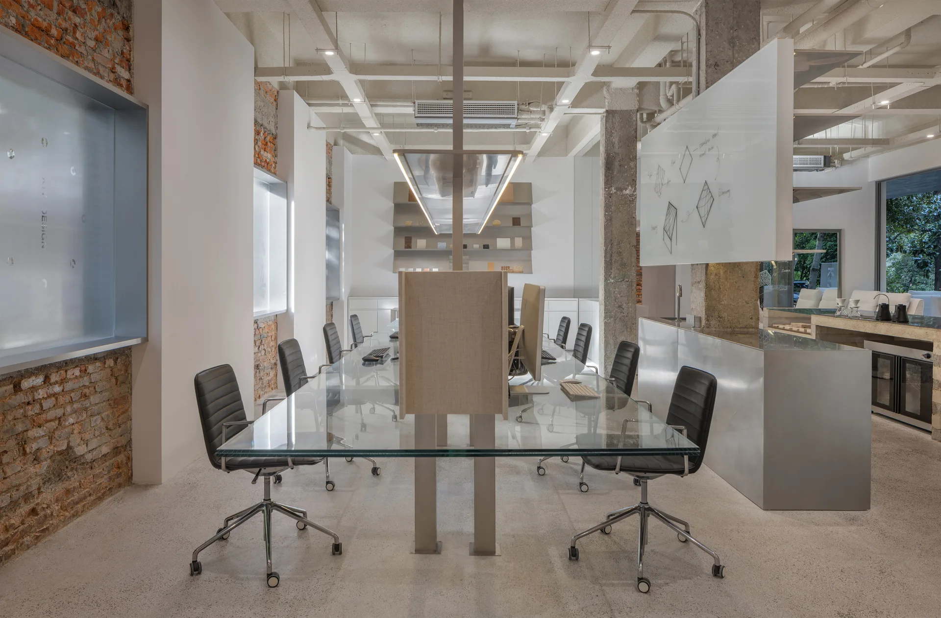 An industrial office features a long glass-top desk lined with black swivel chairs, set against exposed brick walls and concrete columns under a waffle ceiling.
