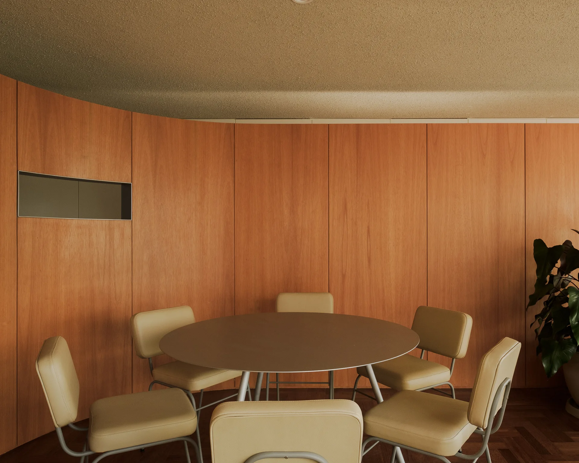 A round dining table surrounded by beige chairs is positioned against a curved wood-paneled wall on parquet flooring.