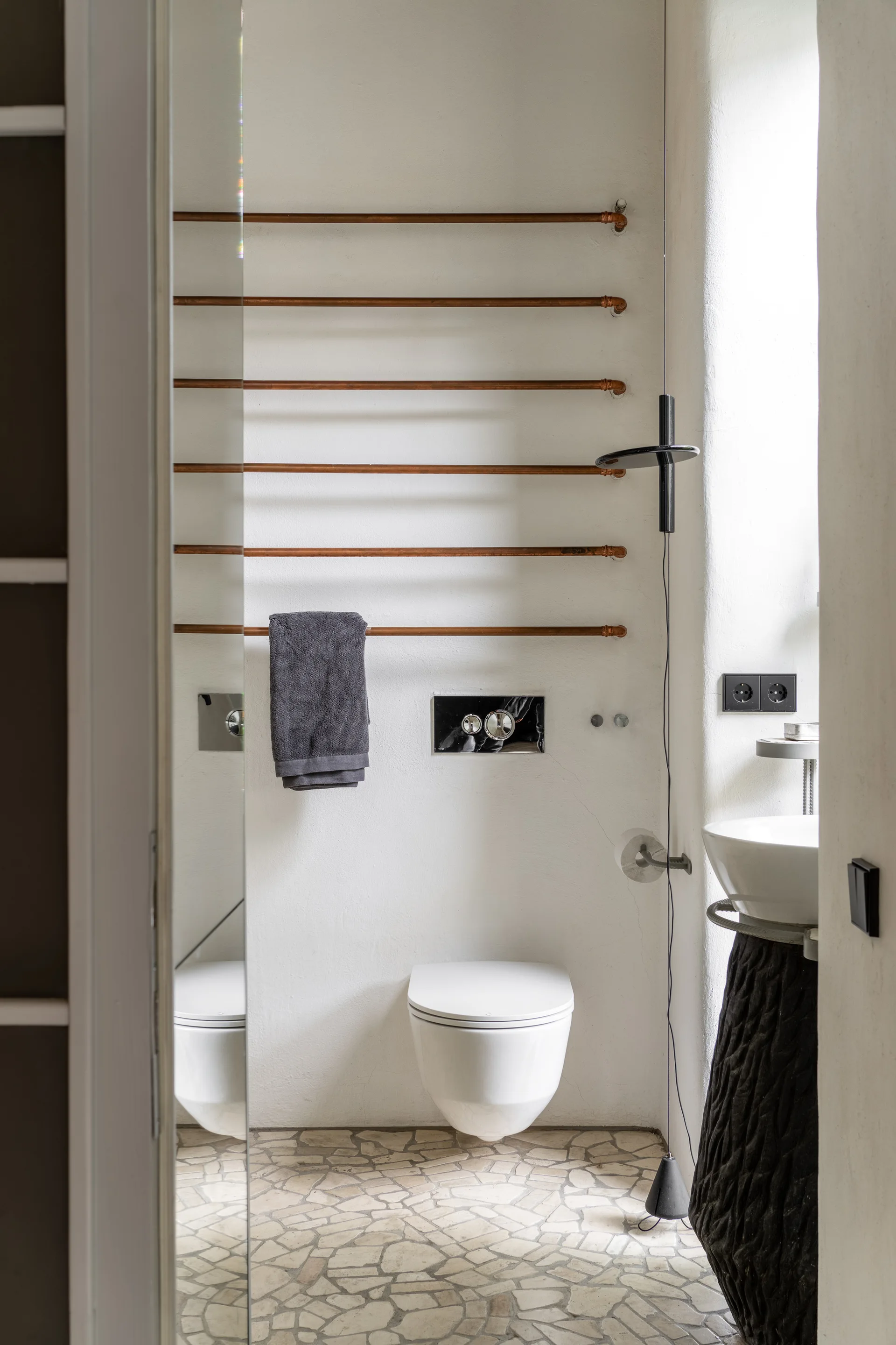 A minimalist bathroom featuring a wall-mounted toilet, several horizontal copper pipe towel rails, and a stone mosaic floor.