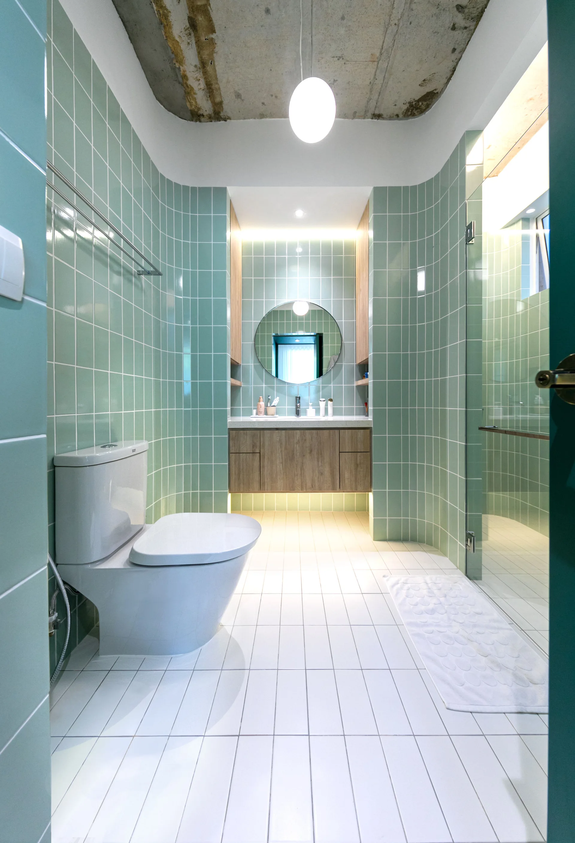 A modern bathroom features mint green tiled walls with curved corners, a floating wood vanity, and an exposed concrete ceiling.