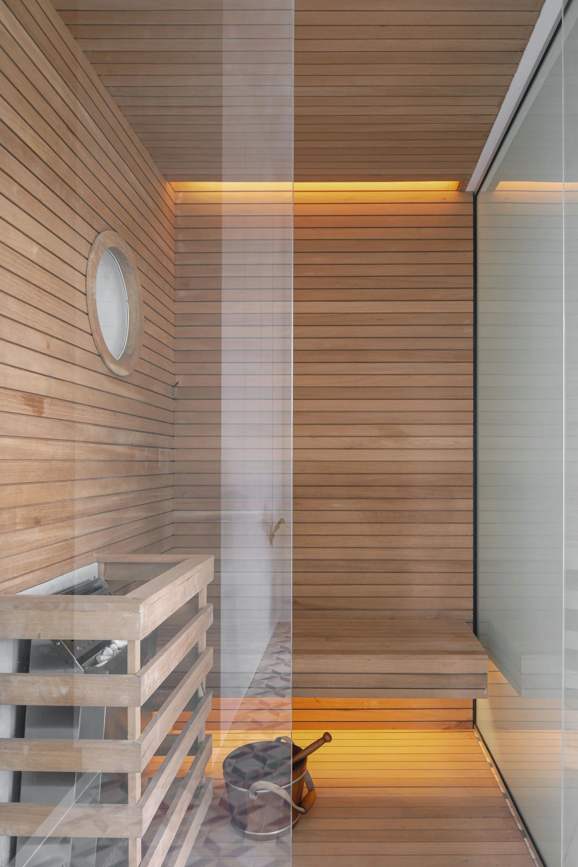 A modern wood-slat sauna interior features a built-in bench, a circular porthole window, and warm, indirect cove lighting behind a glass partition.