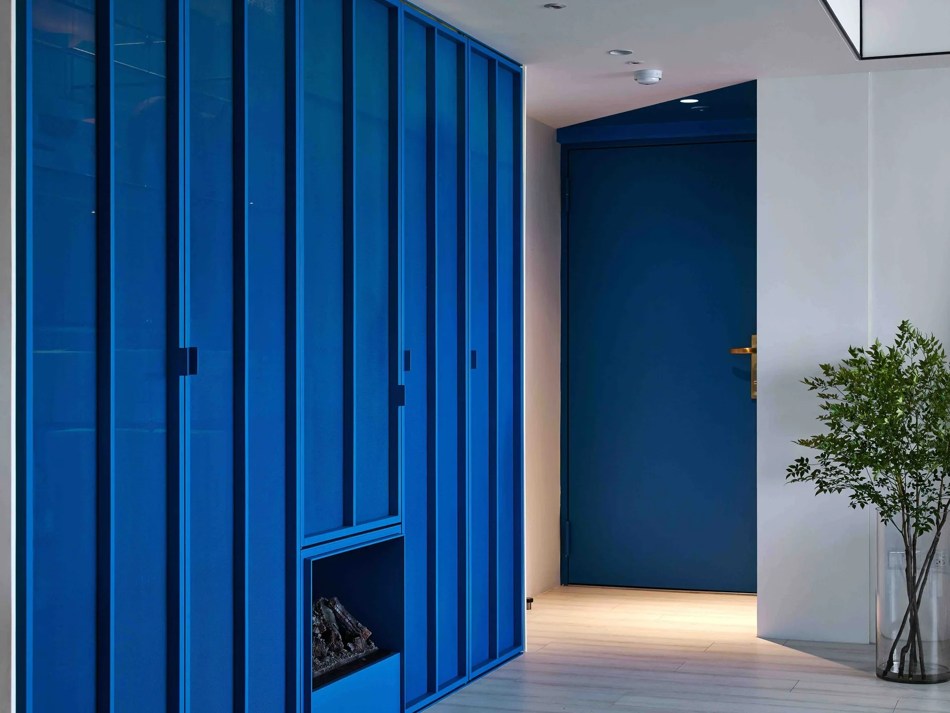 Vibrant blue slatted cabinetry with a small recessed fireplace lines a hallway next to a matching blue door and a clear vase.