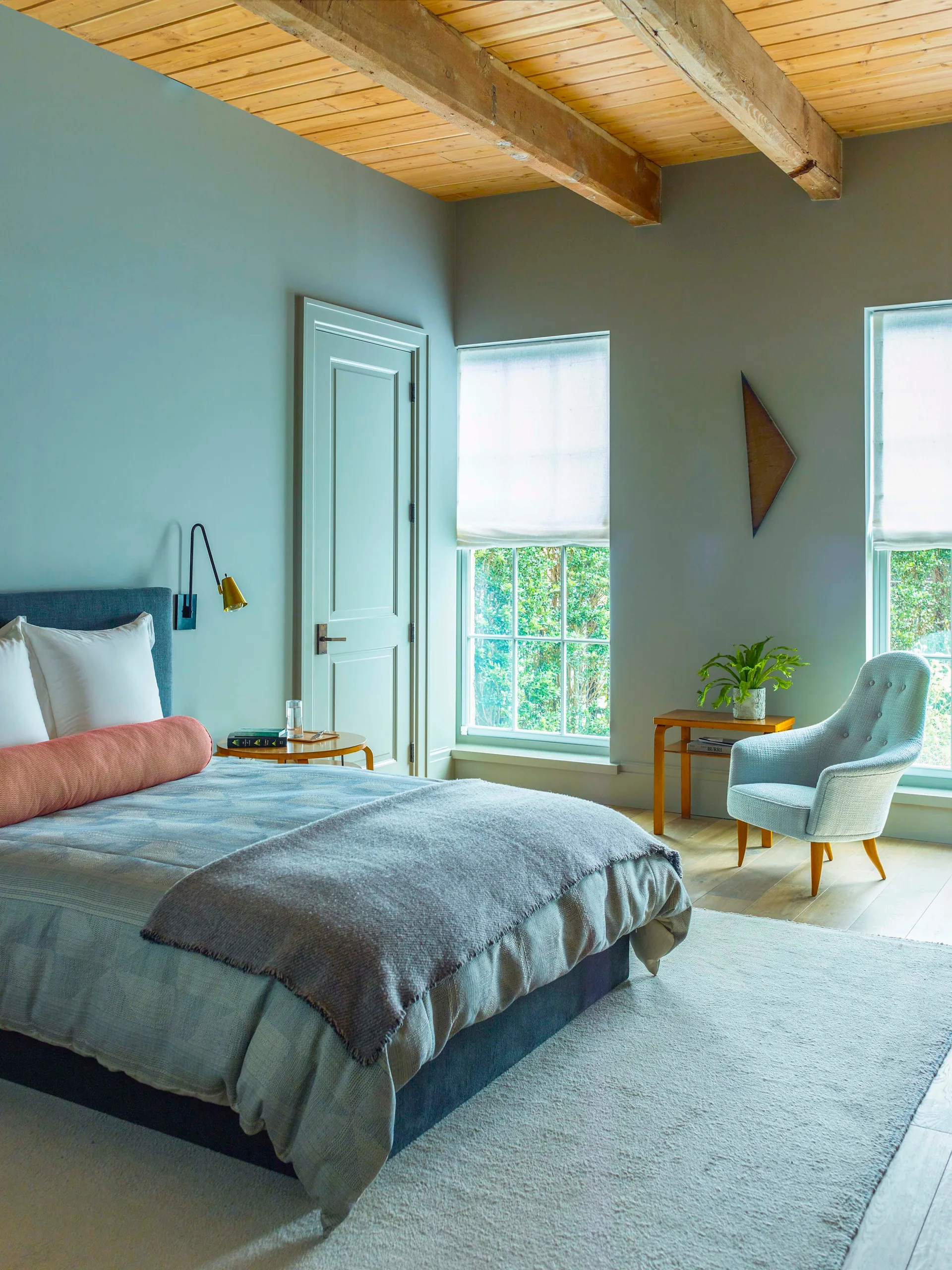 A bedroom with light blue walls, exposed wood ceiling beams, a bed with gray bedding, and a tufted armchair near two windows.