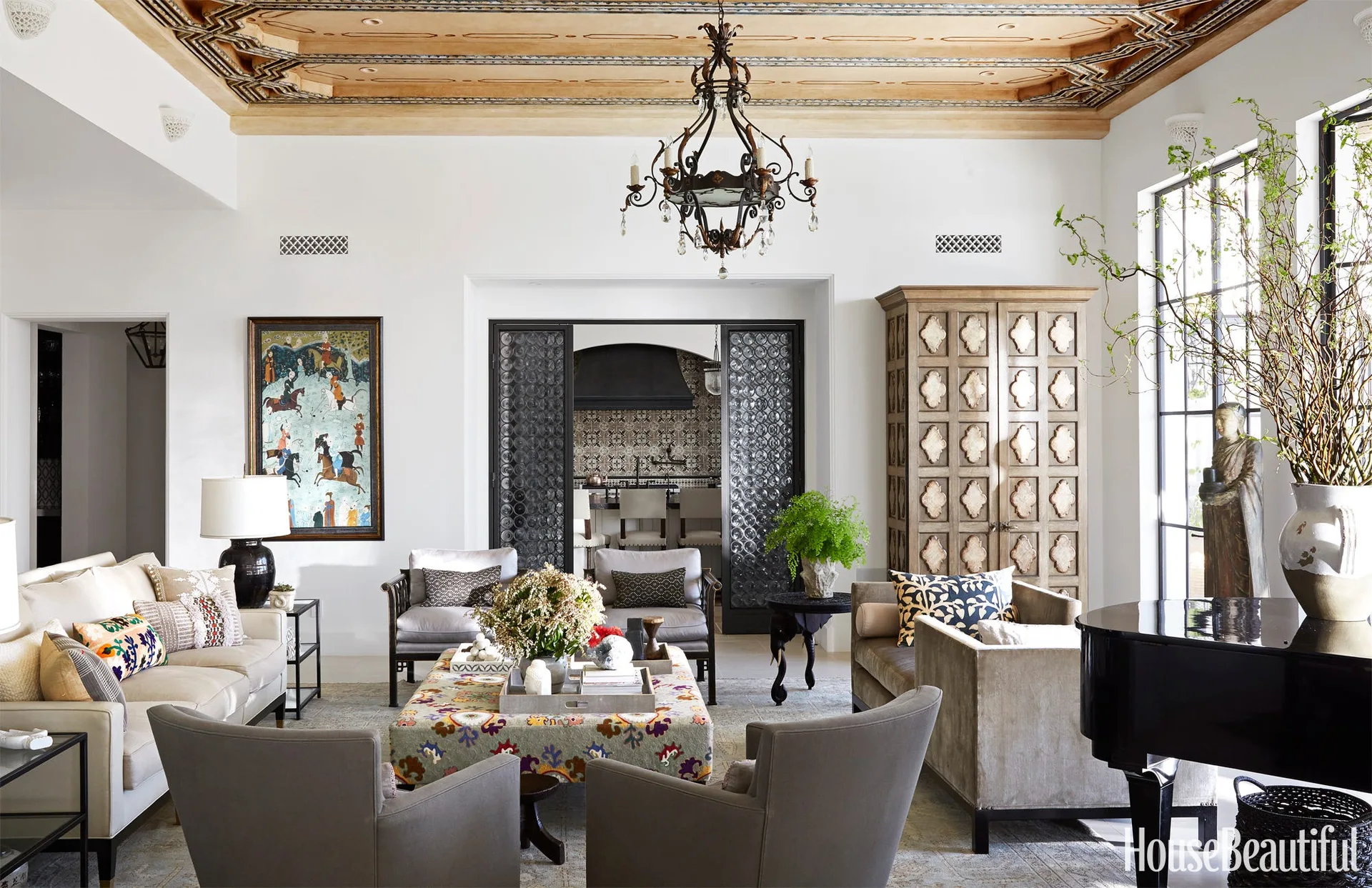 An eclectic living room features white walls, a detailed carved wood ceiling, a large patterned ottoman coffee table, and upholstered seating.