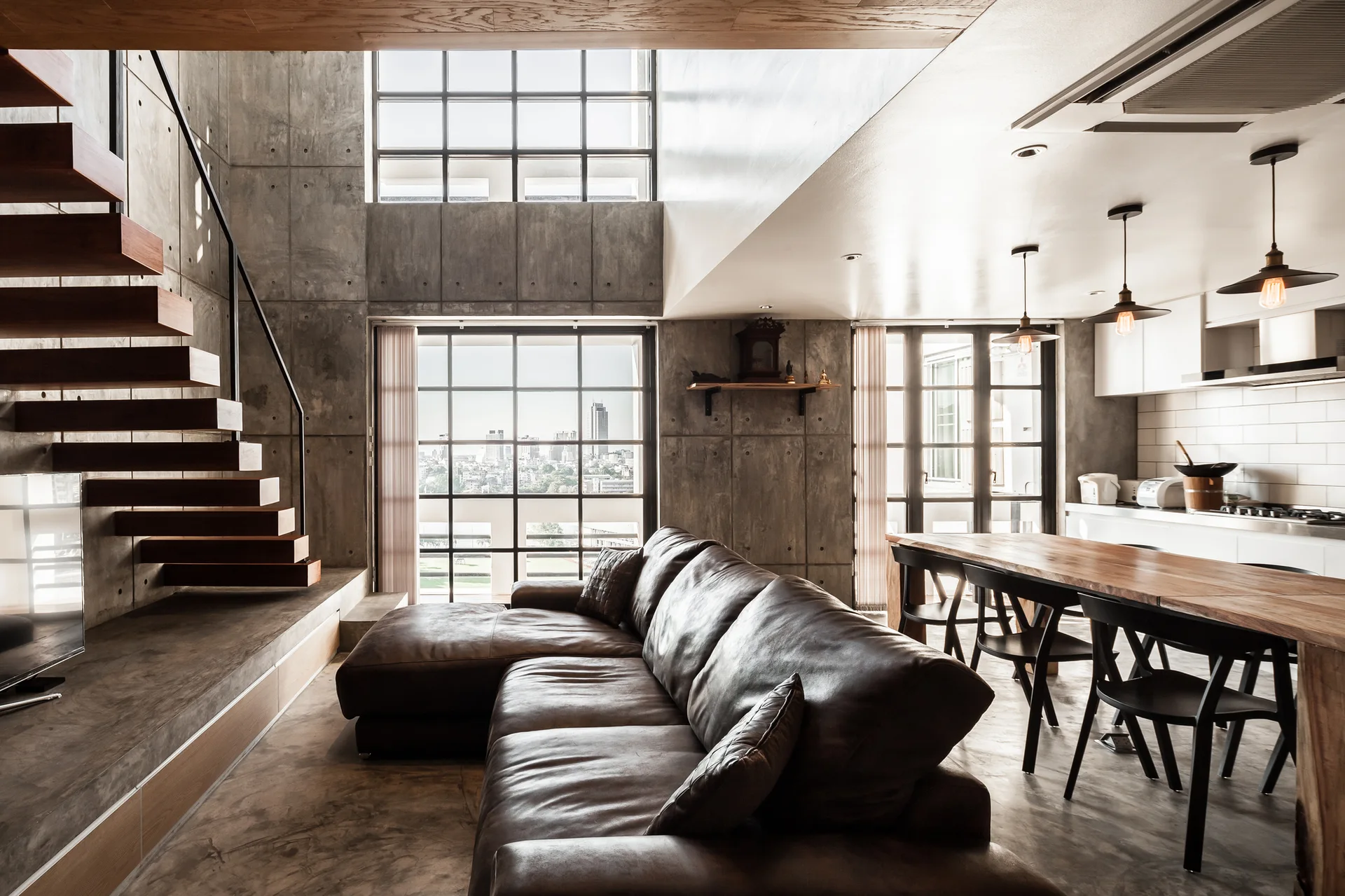 An industrial loft living room with a brown leather sectional sofa, raw concrete walls, and a floating wooden staircase.