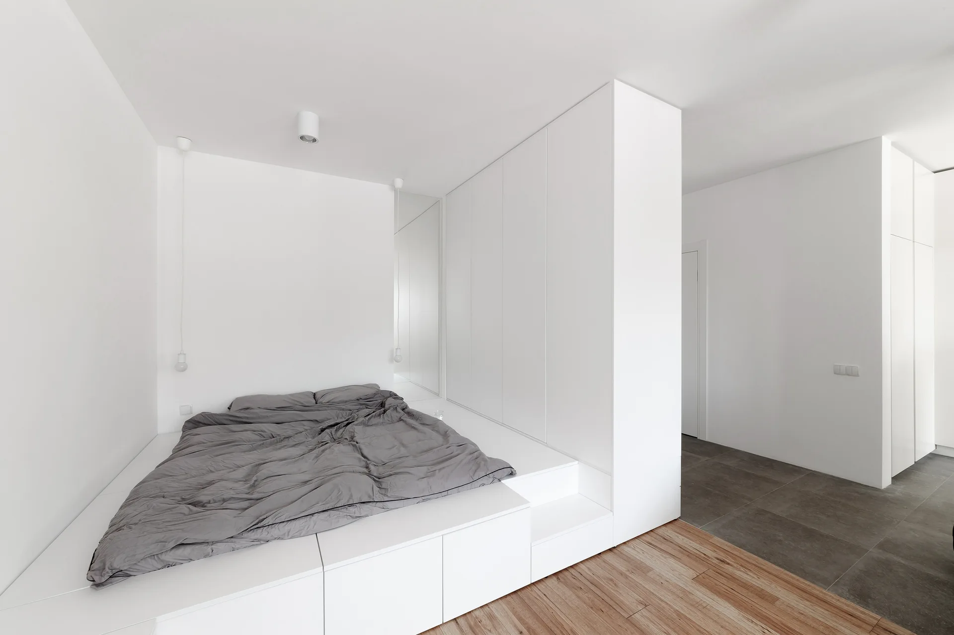 A minimalist white bedroom features a built-in platform bed with grey bedding next to tall integrated floor-to-ceiling wardrobe cabinets.