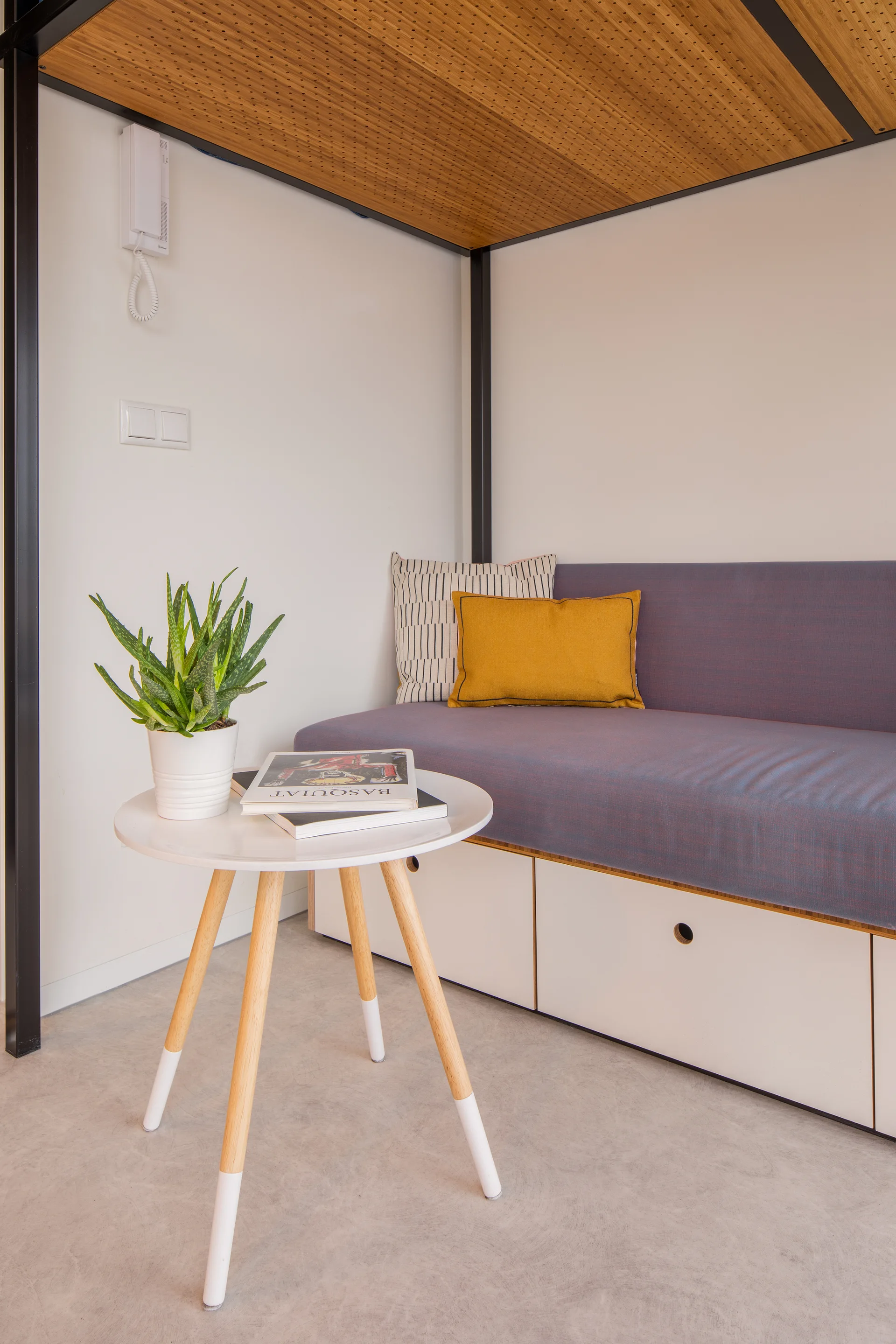 A modern built-in daybed with grey upholstery and storage drawers sits beneath a perforated wood ceiling next to a round white tripod side table.