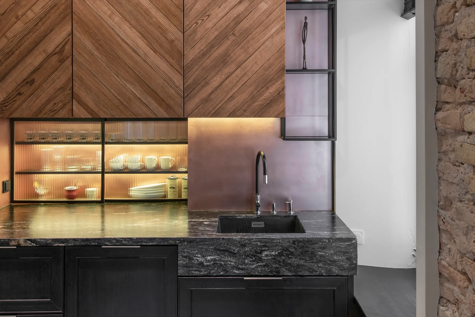 A kitchen sink area features chevron-patterned wood cabinets, a copper backsplash, a black stone countertop, and an illuminated fluted glass cabinet.