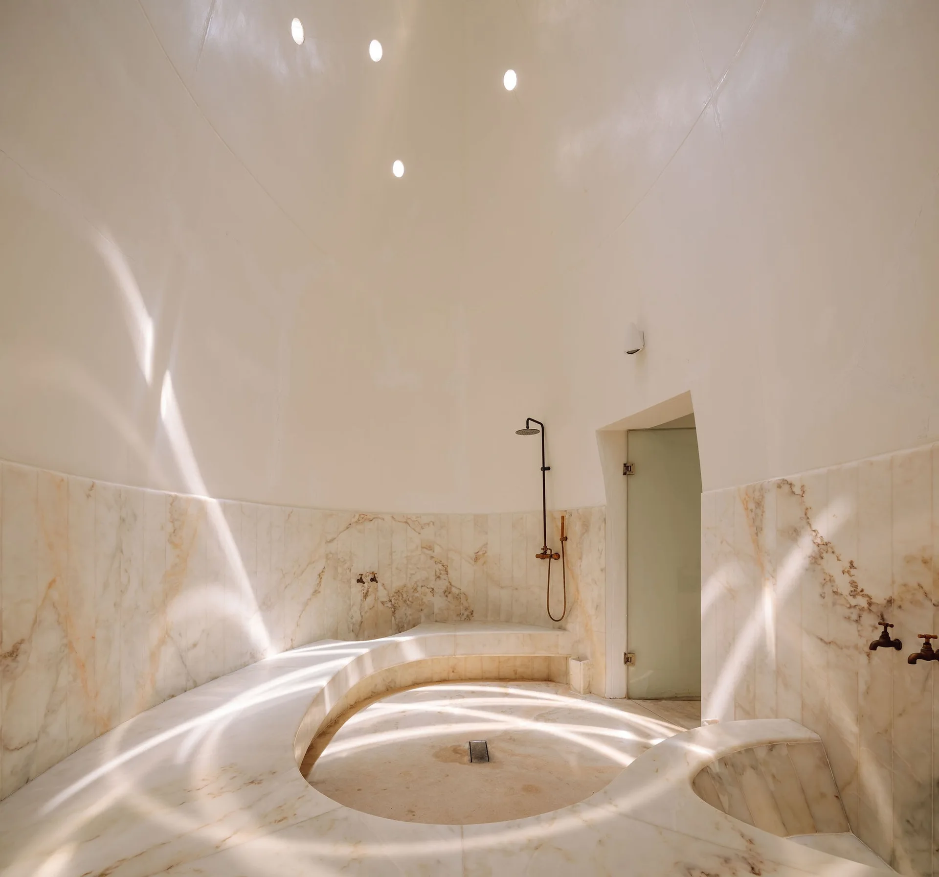 A circular spa room featuring a curved marble bench, matching marble wall cladding, a shower fixture, and a smooth domed ceiling with recessed lights.