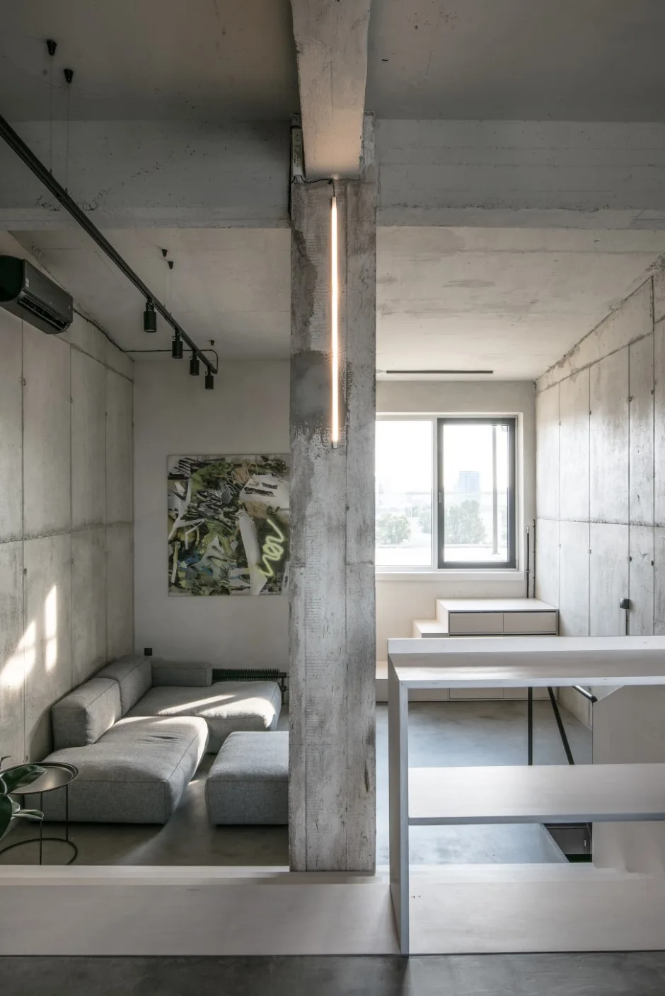 A minimalist living room featuring raw concrete walls, a low-profile grey sofa, and a central concrete column with an integrated vertical strip light.
