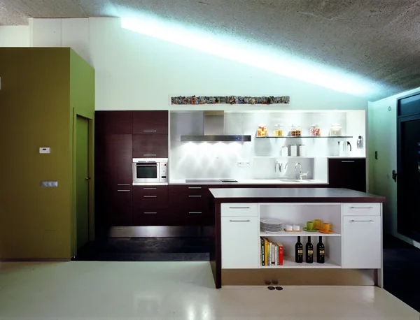 A modern kitchen features dark brown cabinetry, a white island with open shelving, and a textured sloped ceiling illuminated by bright cove lighting.