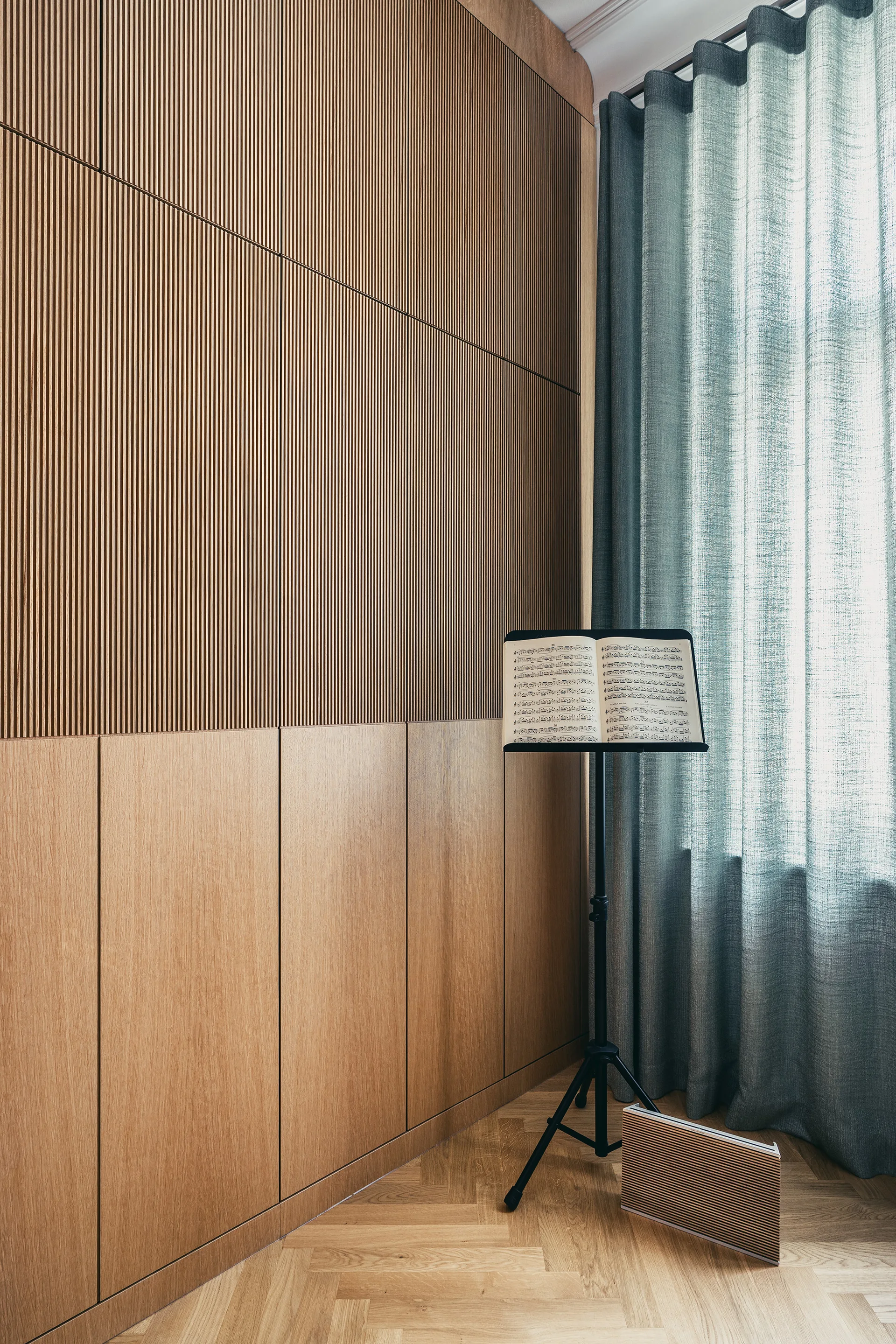A room corner features ribbed wood wall paneling above flat cabinets, herringbone parquet flooring, a black music stand, and blue-grey curtains.