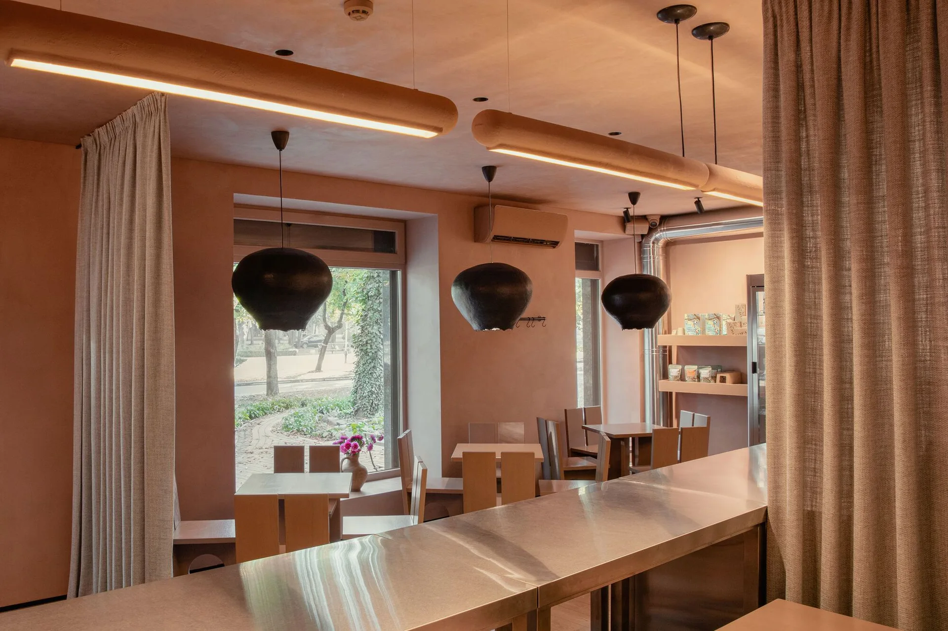 A warm pinkish-beige cafe dining area featuring wooden tables, black organic-shaped pendant lights, and a long brushed stainless steel counter.