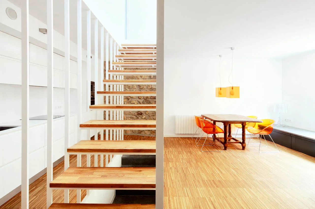 A modern open-plan dining room features a floating wood staircase with white vertical railings, a rustic dining table, and translucent orange chairs.