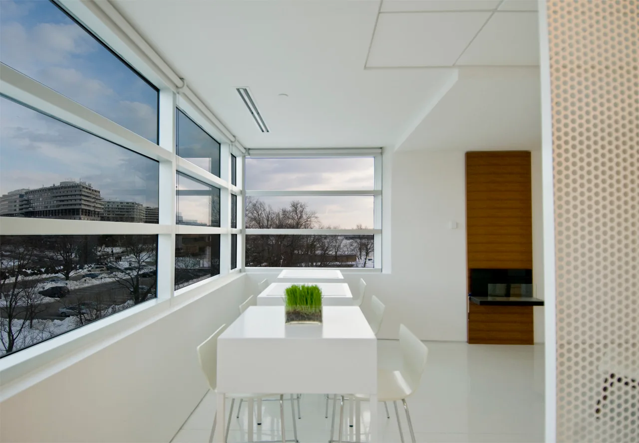 A bright minimalist dining area featuring white tables and chairs, glossy white flooring, and large grid windows overlooking a snowy landscape.