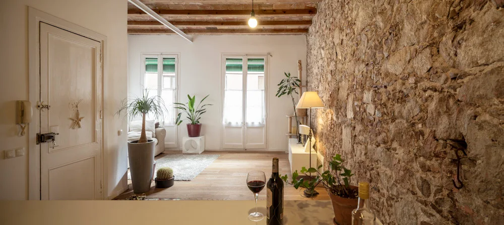 A rustic living room with a textured stone wall, exposed wood ceiling beams, a light gray rug, and potted plants next to French doors.