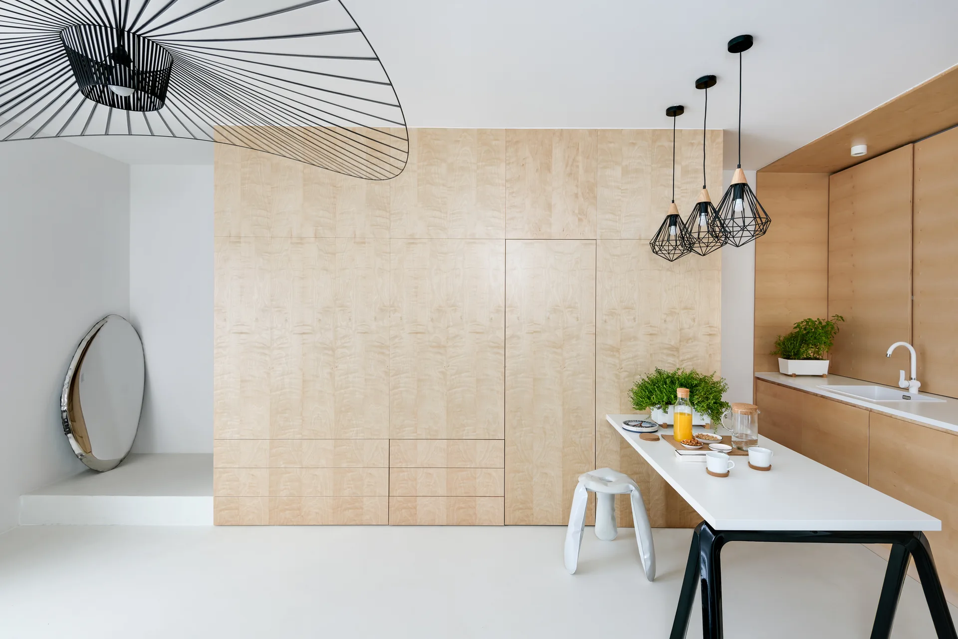 A minimalist dining space features a seamless light wood storage wall, a white table with a white stool, and black wire-frame pendant lights.